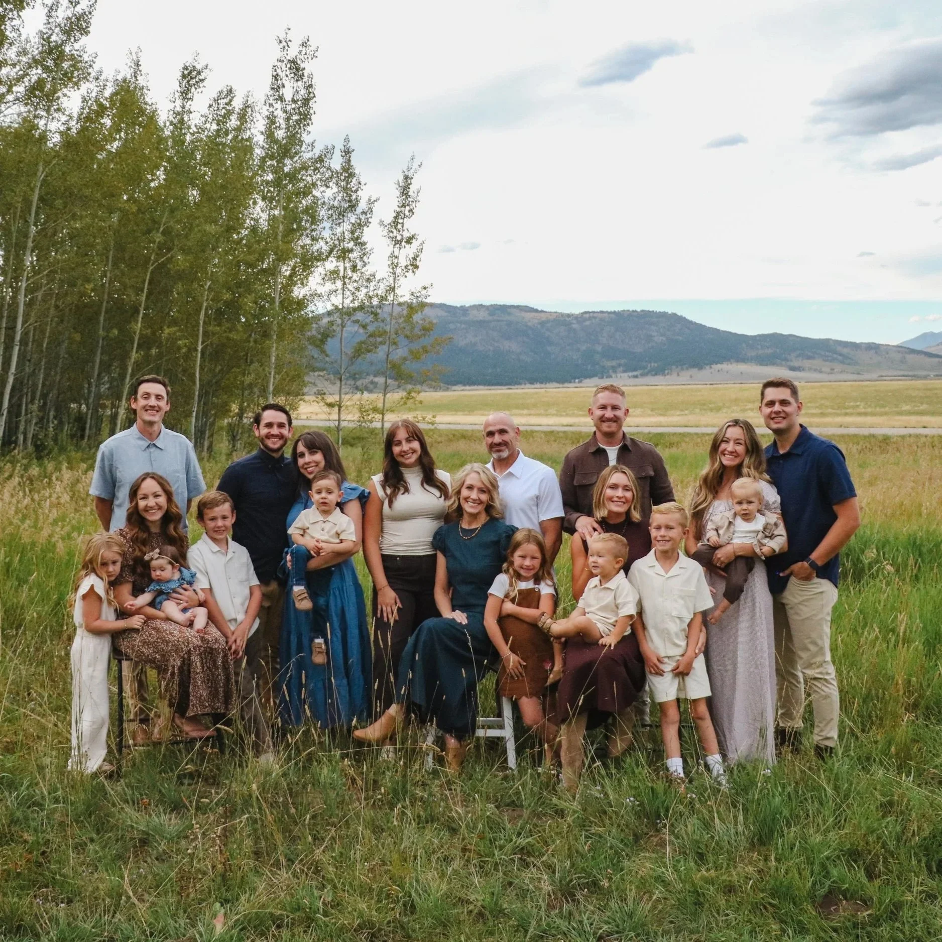 A group of adults and children standing outdoors in a grassy field with trees and mountains in the background, looking at the camera and smiling.