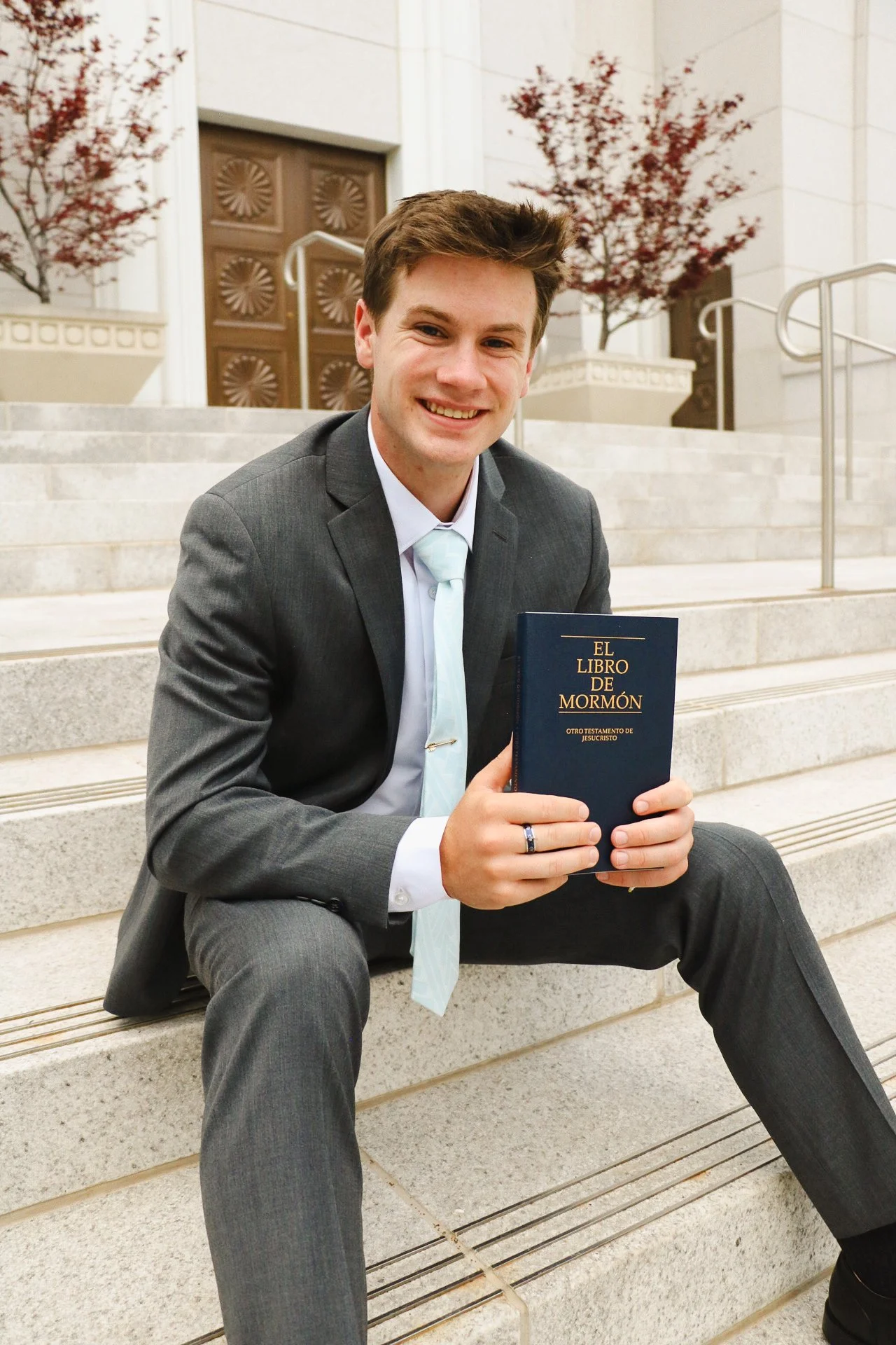 A young man in a gray suit and white shirt sitting on outdoor stairs, smiling, holding a book titled 'El Libro de Mormón' in Spanish.