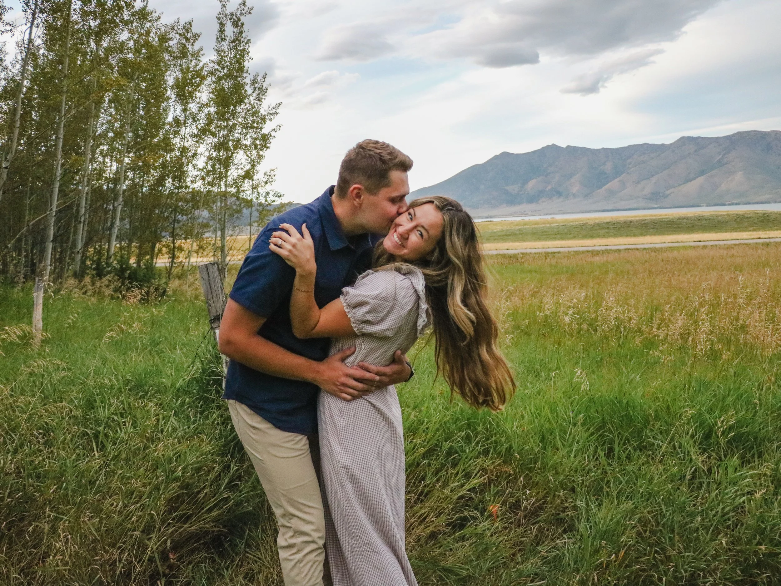 A couple embracing outdoors in a grassy field near trees with mountains and a cloudy sky in the background.