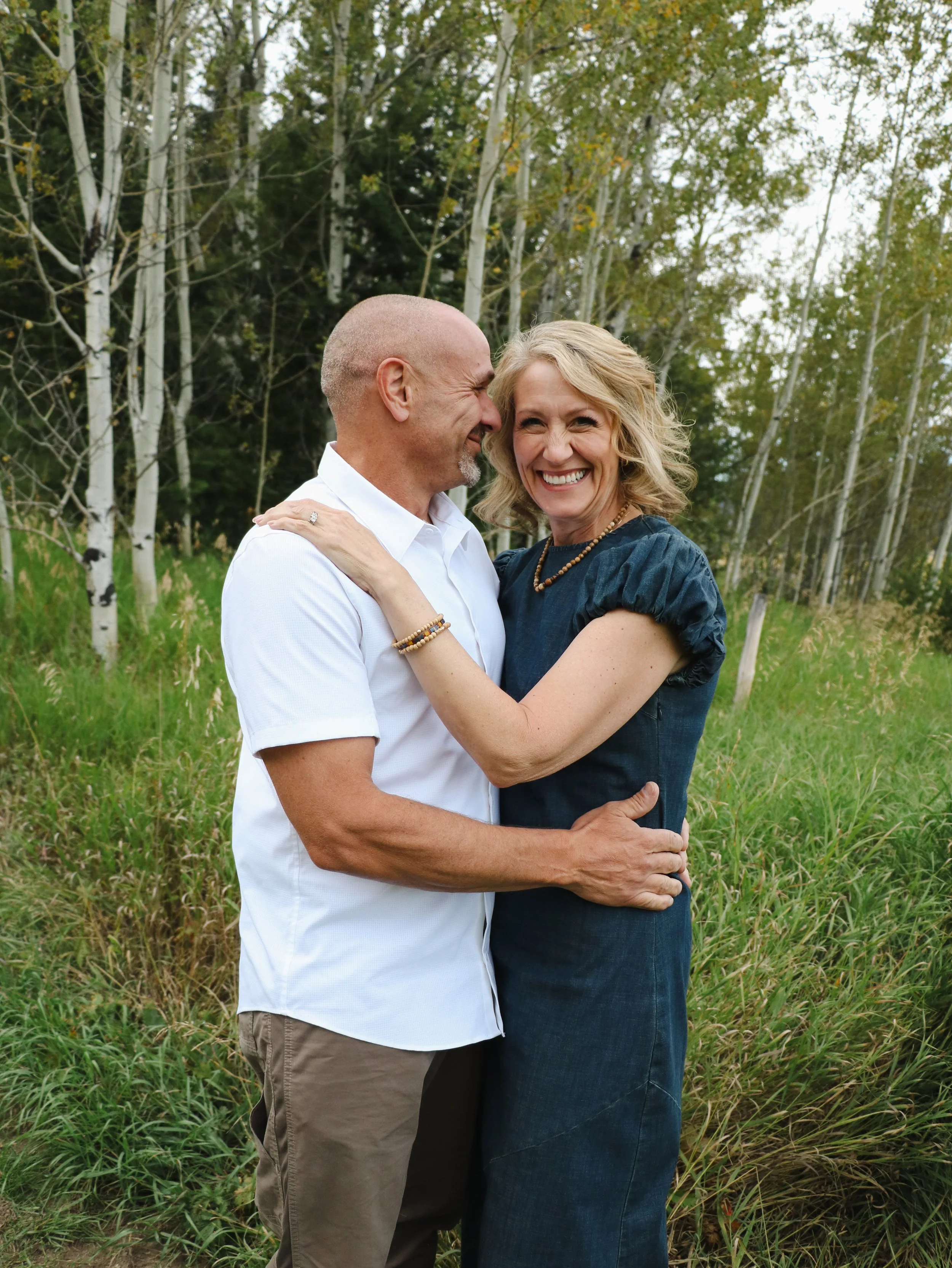 A happy couple embraces outdoors in a grassy area with trees in the background.