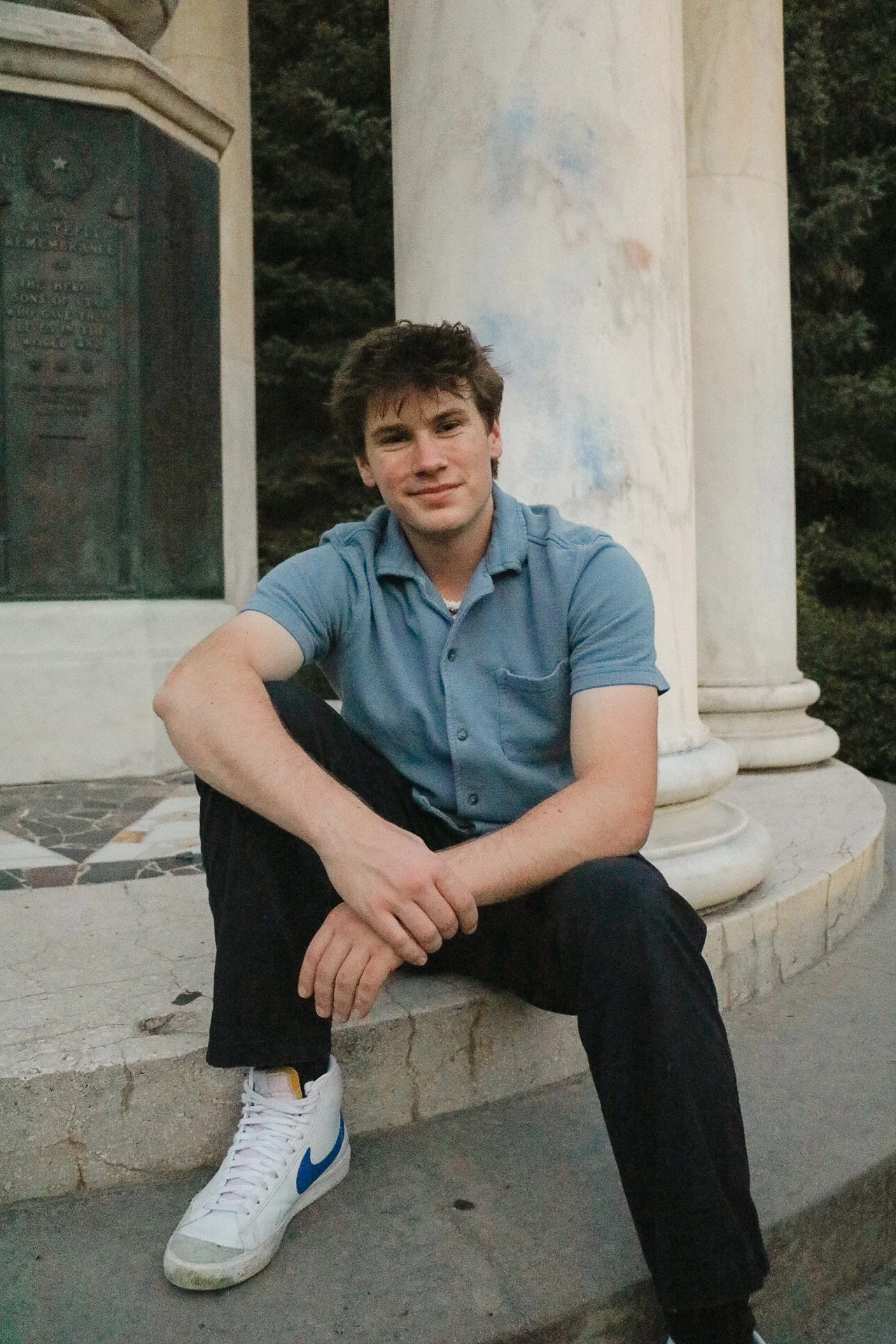 Young man with short brown hair and fair skin sitting on a marble bench in front of white columns, wearing a light blue polo shirt, black pants, and white sneakers with blue and yellow accents.