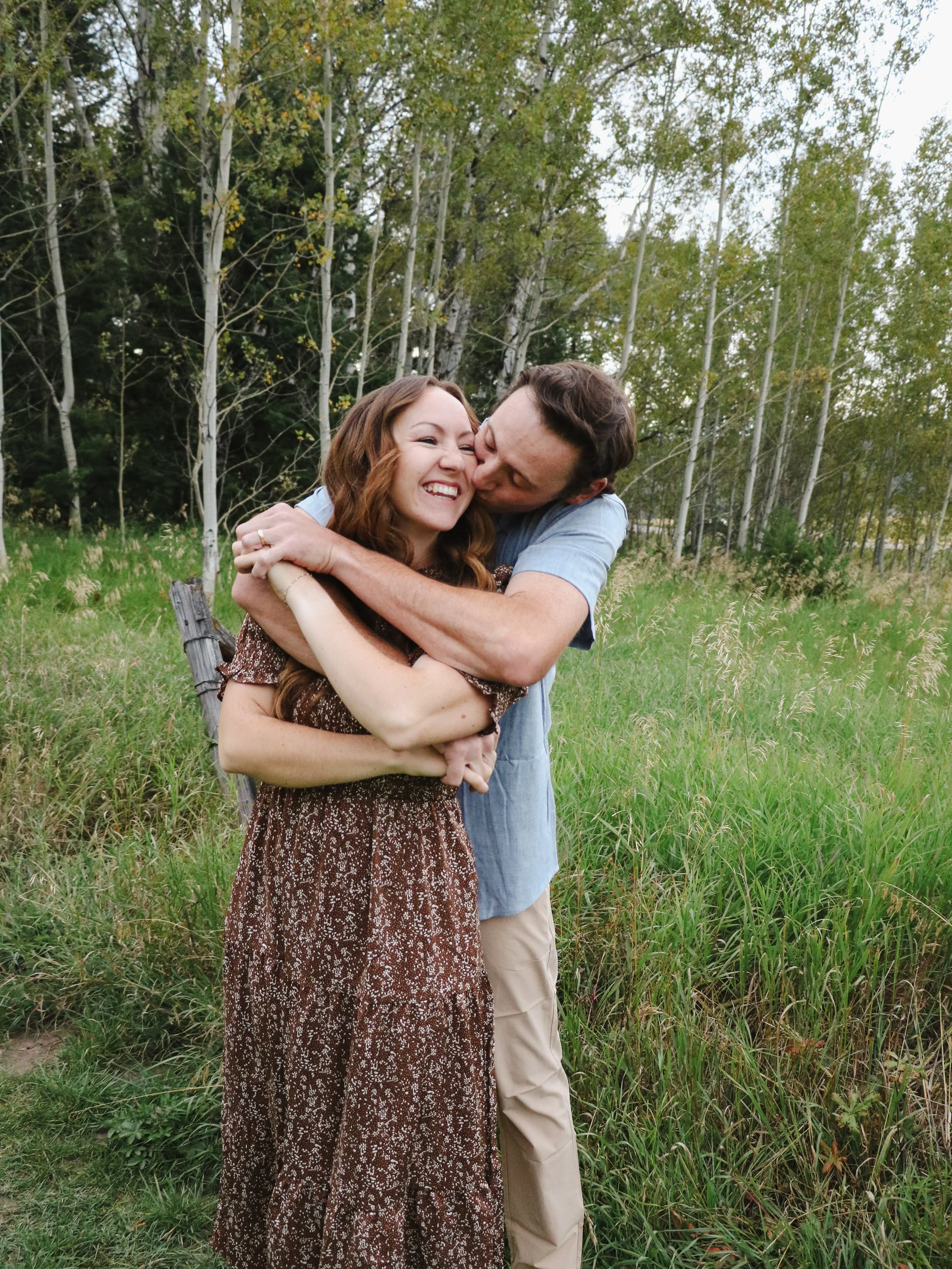 A happy couple hugging outdoors in a field with green grass and tall trees.
