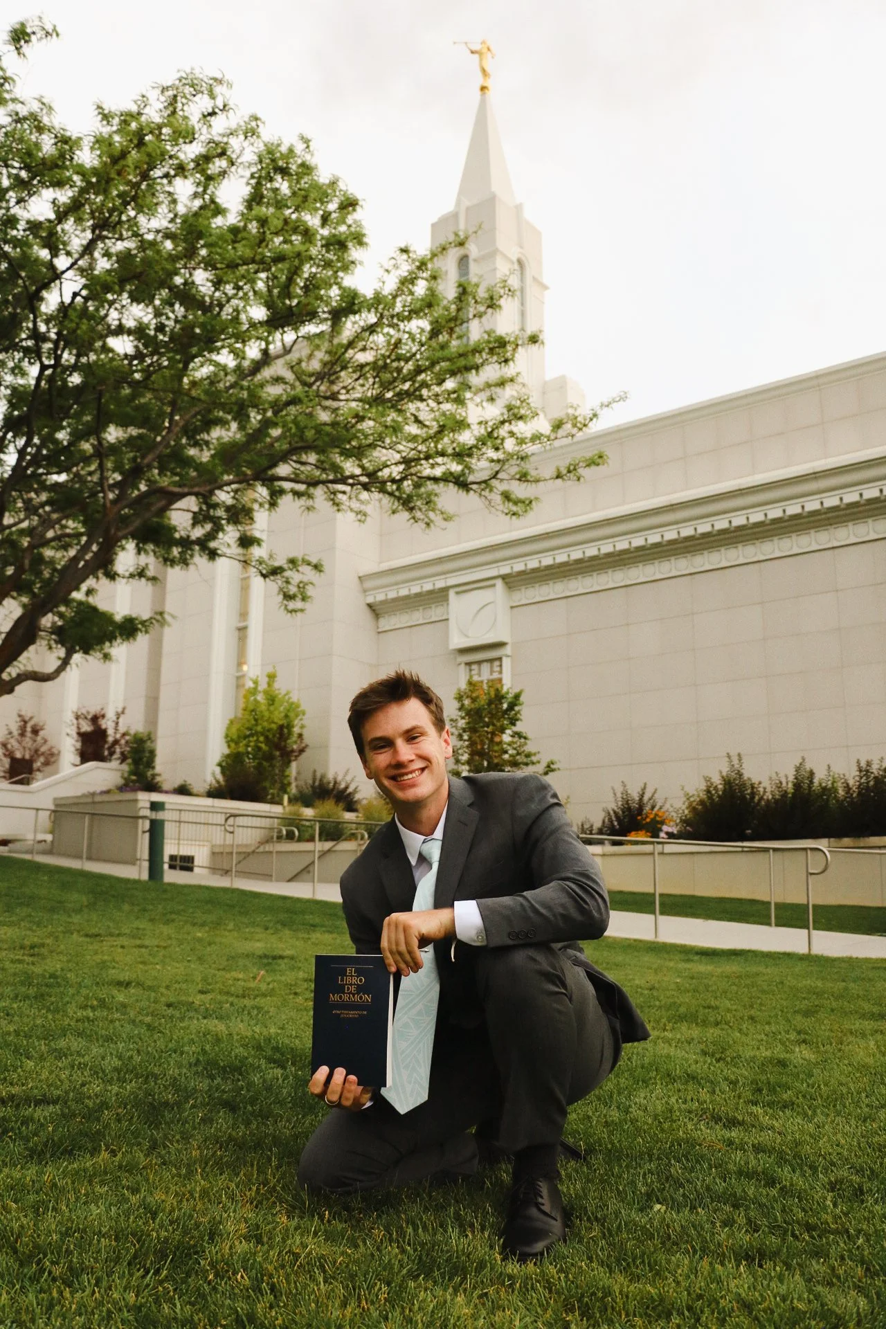 A young man in a suit kneeling on the grass outside a church, holding a book titled 'El Libro de Mormón' and smiling at the camera.