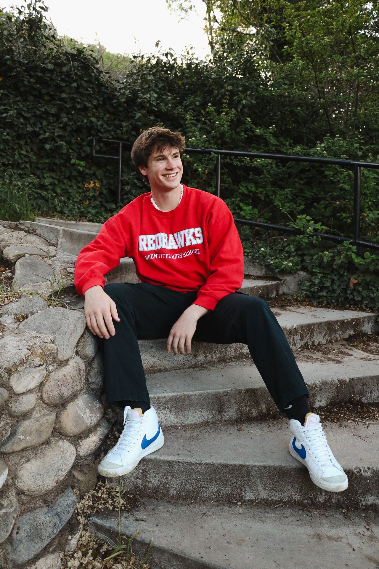 A young man sitting on outdoor concrete stairs, wearing a red sweatshirt with "Redhawks Bountiful High School" printed on it, black pants, and white sneakers with blue Nike swooshes. He is smiling and looking to his left, with greenery and a black me