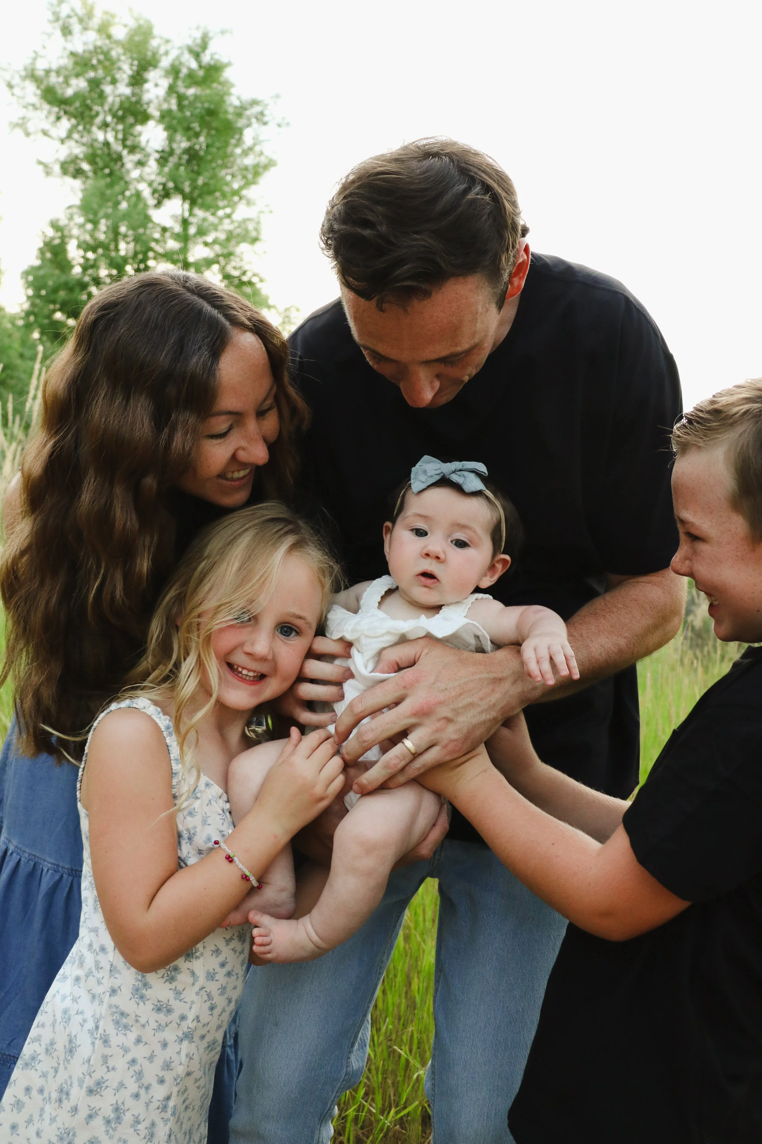 A group of people, including children and adults, gathered outdoors holding a baby, with trees and grass in the background.