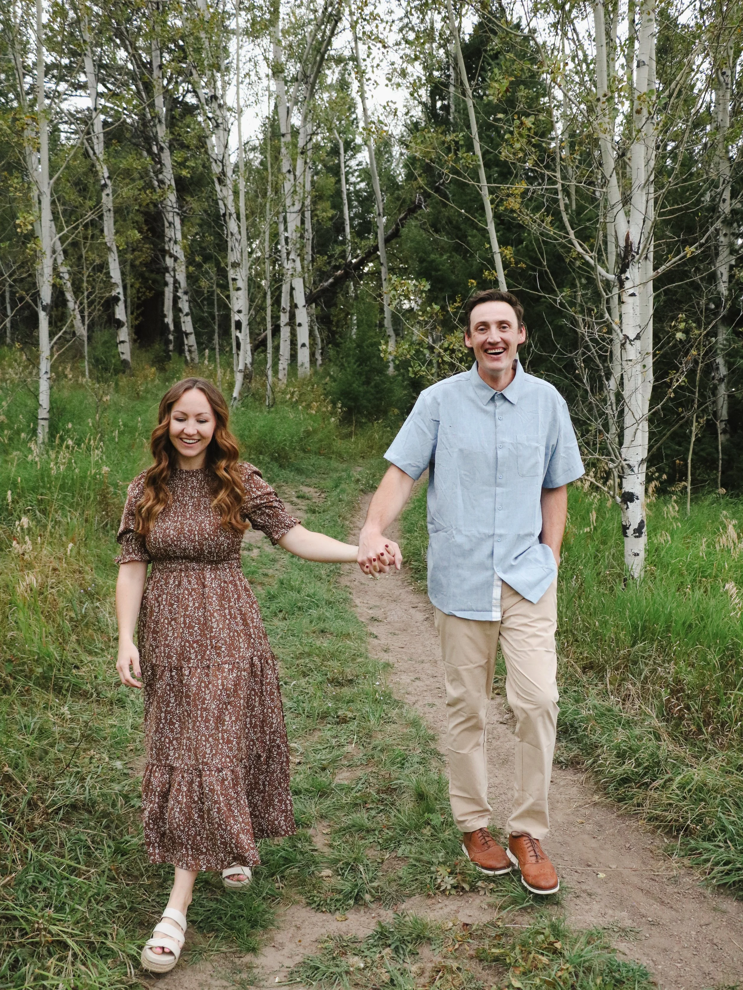 A man and woman holding hands walking on a dirt path through a forest with green grass and trees.