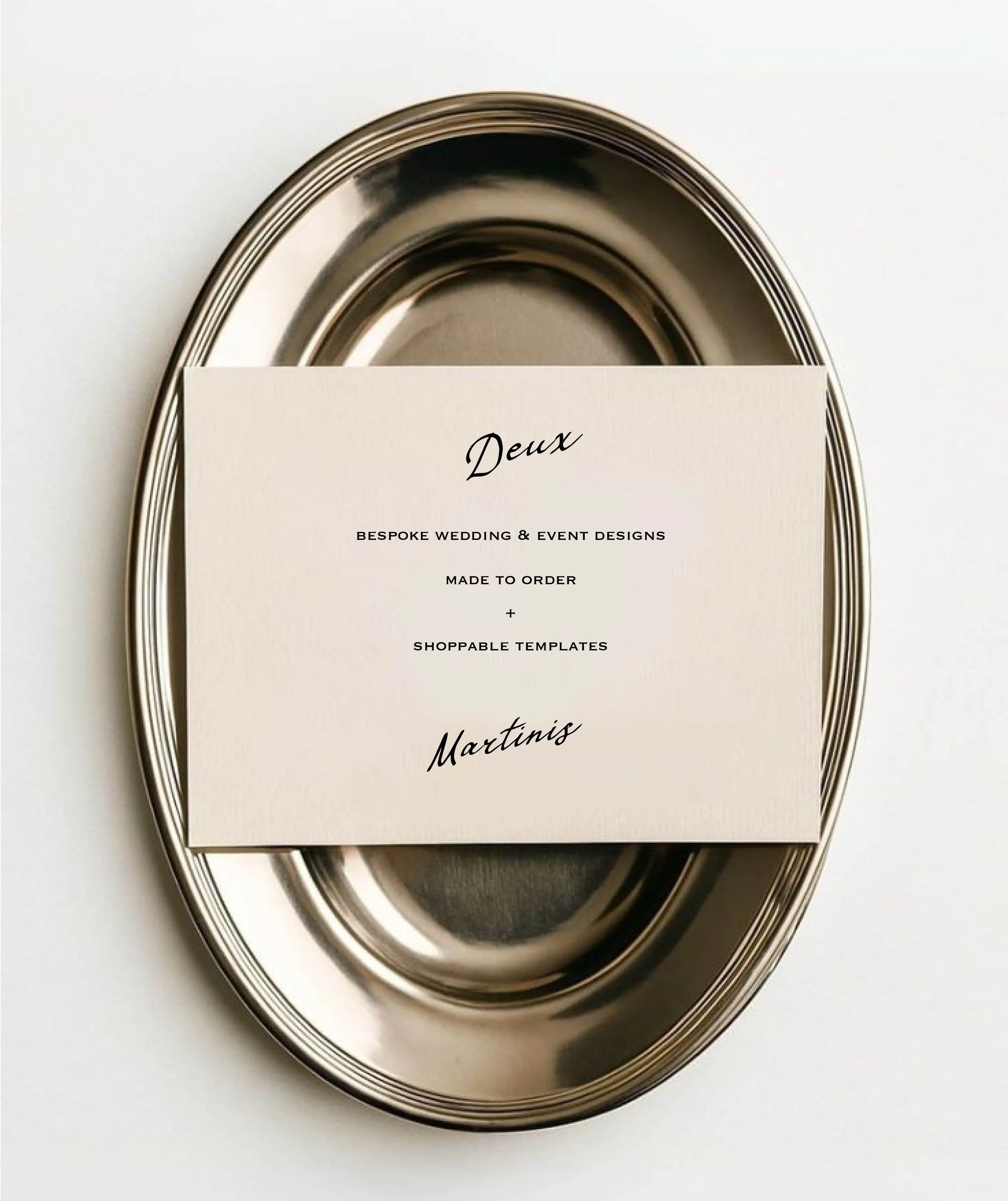 A white card with elegant cursive text reading 'Deux' and additional text below stating 'Bespoke Wedding & Event Designs, Made to Order, Shoppable Templates' partially covering a shiny metal tray or lid, with the tray's slightly capped edge visible.