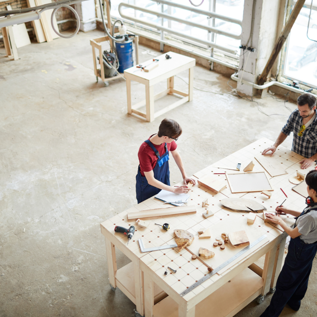 People working on woodworking projects in a woodworking workshop with tools and wooden pieces on a large worktable.