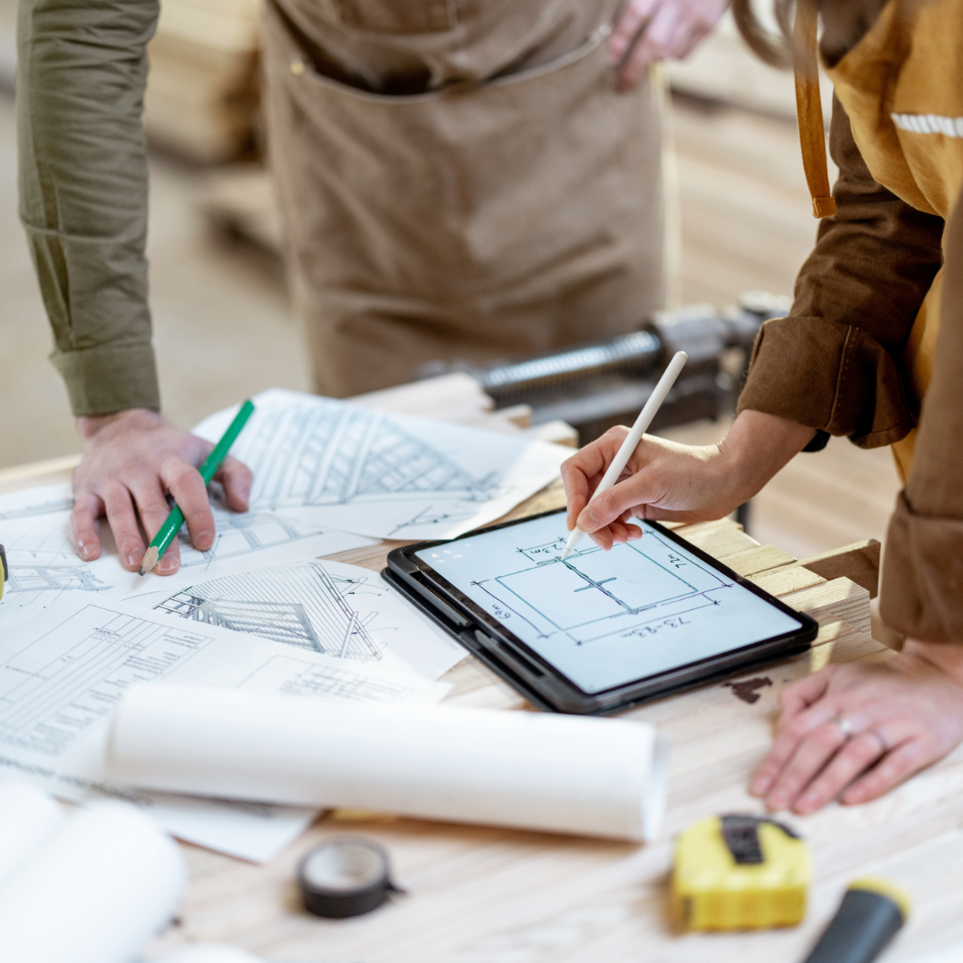 Two people working on architectural plans at a construction site, with one using a tablet to draw and the other examining printed blueprints.