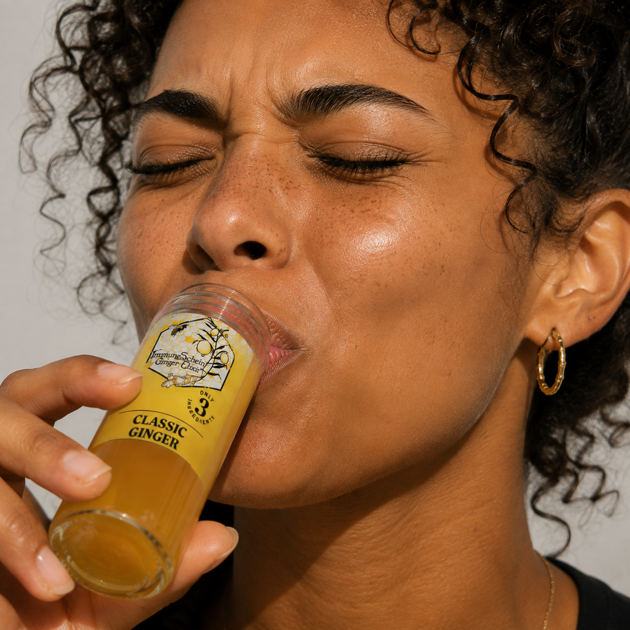 Close-up of a woman with curly hair and earrings drinking ginger shot from a small glass labeled 'Classic Ginger' with a lemon illustration.