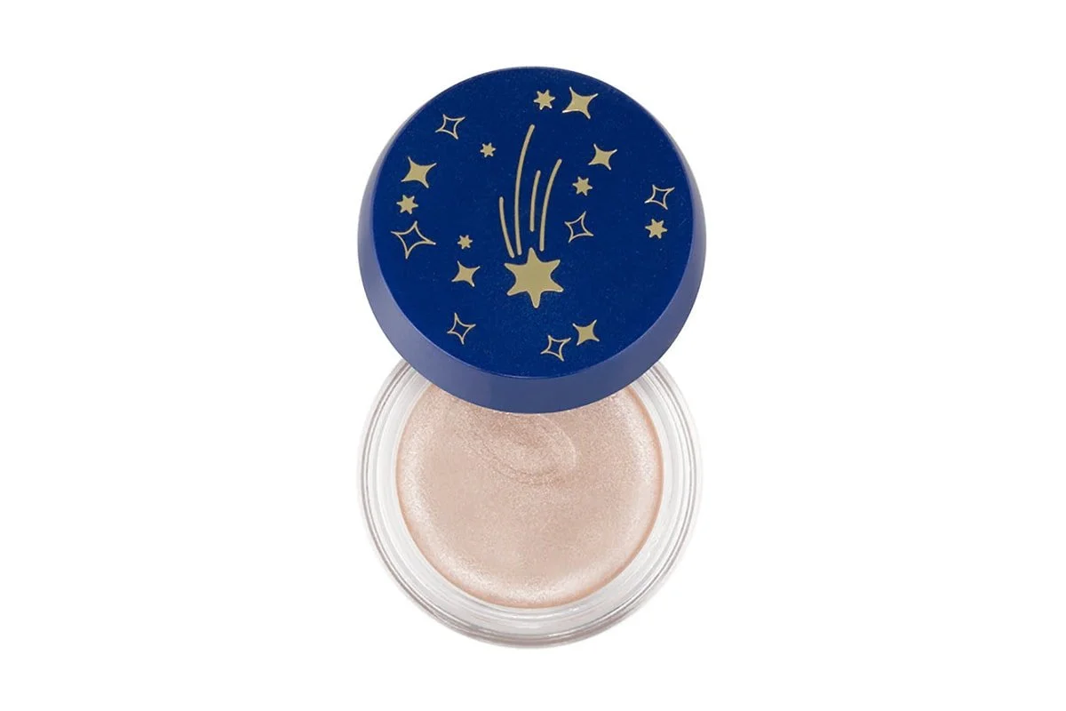 A small round container with a blue lid decorated with gold stars and a shooting star, filled with a shimmery, light-colored cream or gel makeup product.