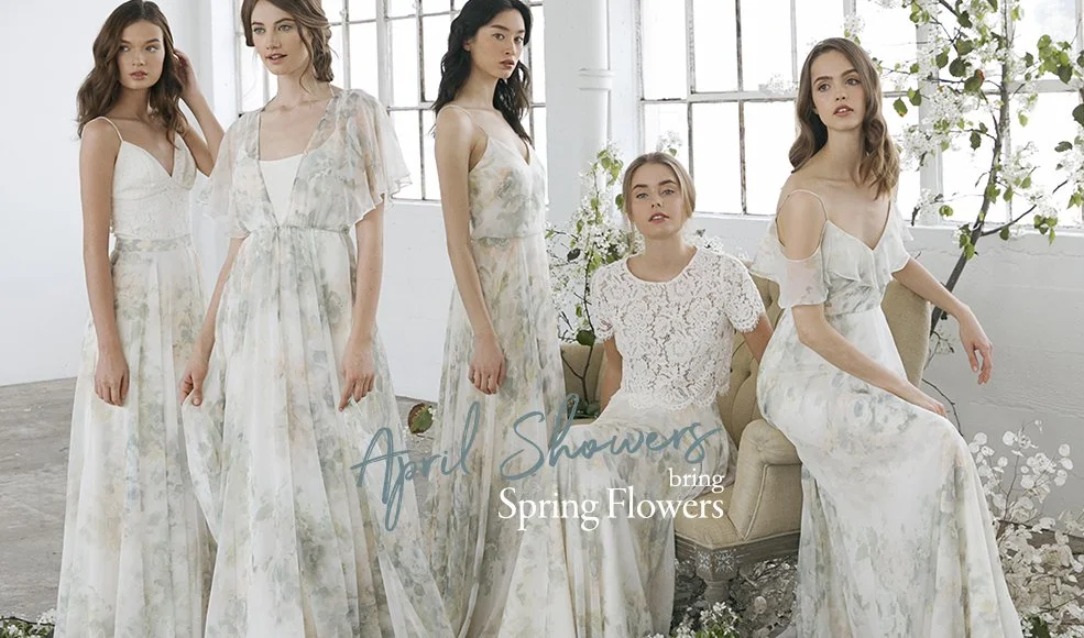 Five young women wearing light-colored, floral-patterned dresses in a bright, airy room with large windows and spring flowers, with text overlay reading 'April Showers bring Spring Flowers.'