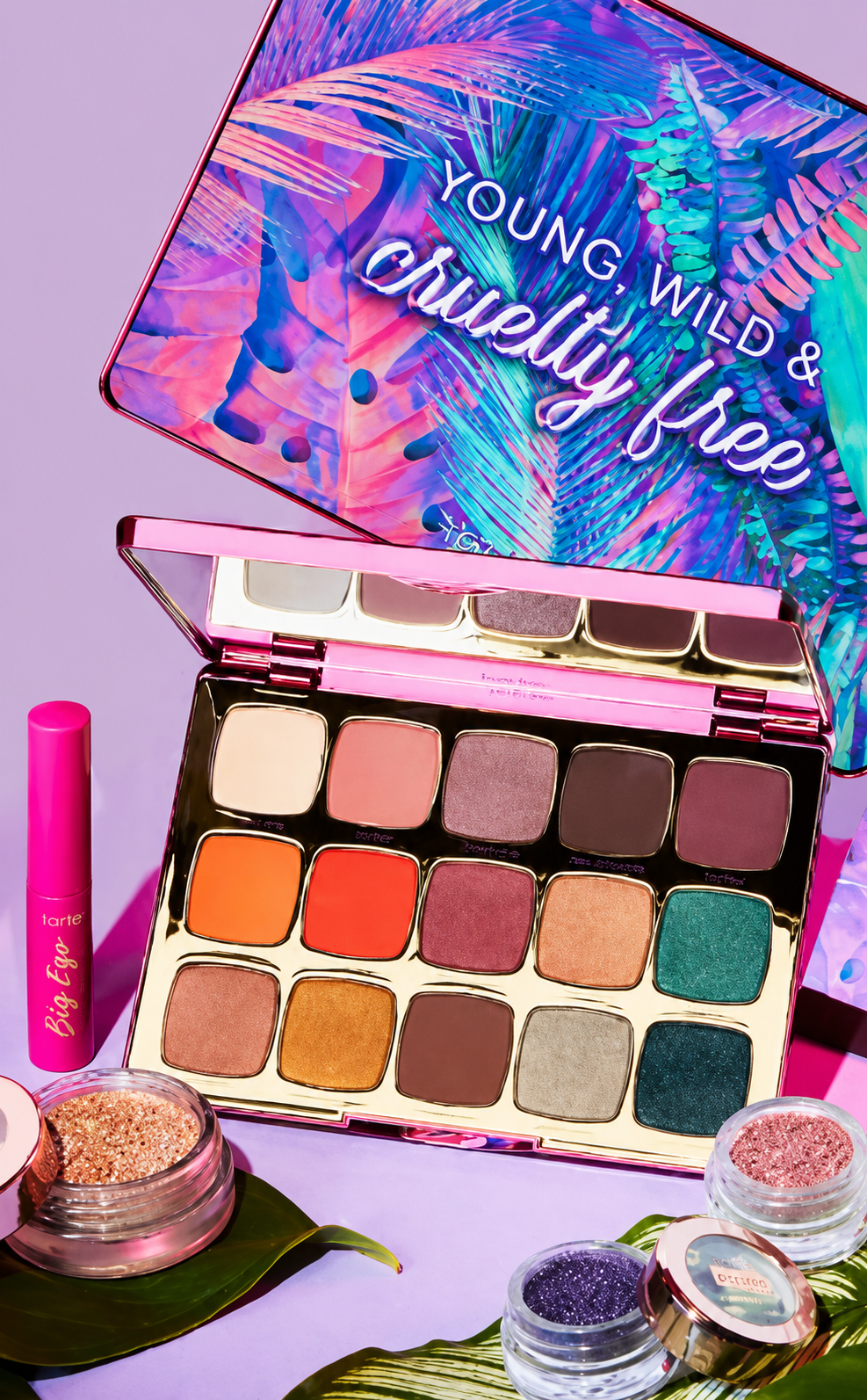 Open eyeshadow palette with colorful shades on a purple surface, surrounded by glitter jars and a pink mascara tube, with a pink and purple tropical-themed message on the lid that says, "Young, Wild & Guilty Free."
