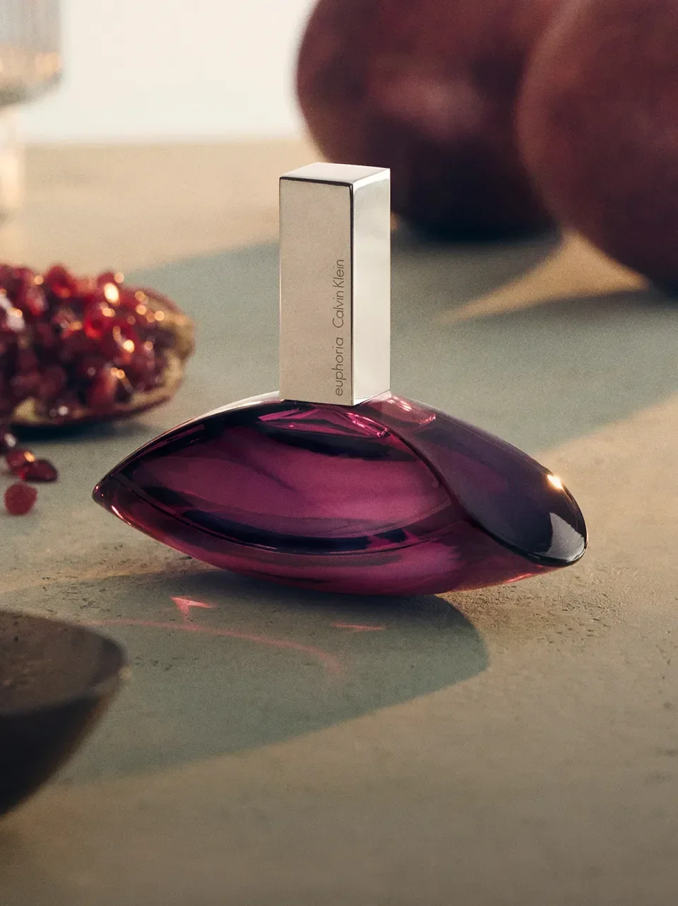 A bottle of Calvin Klein Euphoria perfume placed on a purple glass art piece on a light-colored surface. In the background, there are red gemstones and some brown objects.
