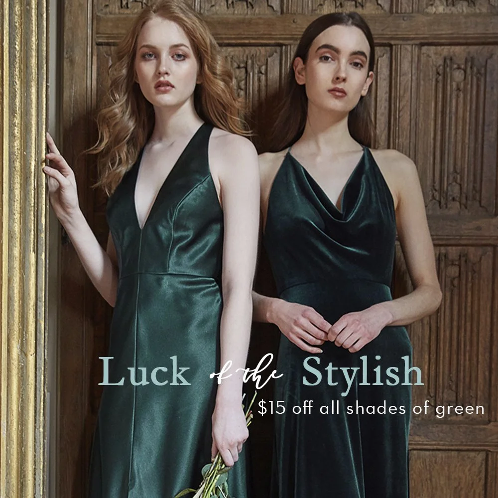 Two women with fair skin and long hair, dressed in elegant dark green satin dresses, standing against a wooden wall. One woman has red hair and the other has dark brown hair, posing for a fashion advertisement.