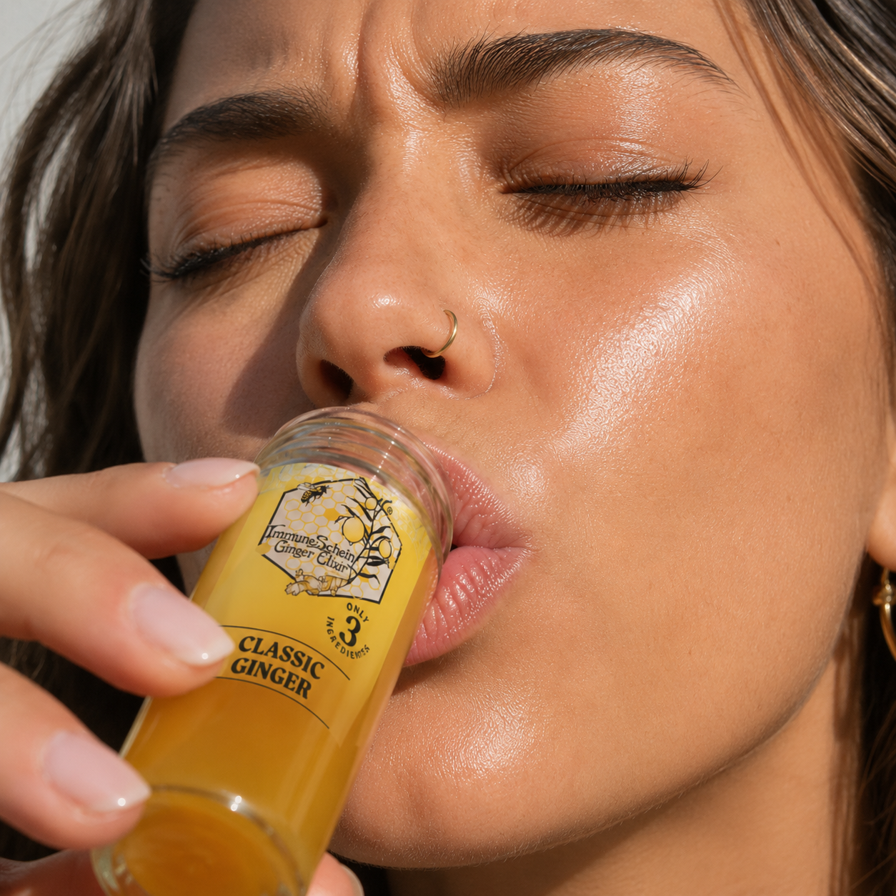 A woman with long hair, a nose ring, and gold earrings is drinking a yellow beverage labeled 'Classic Ginger' from a small glass bottle with a honeycomb and ginger root design.