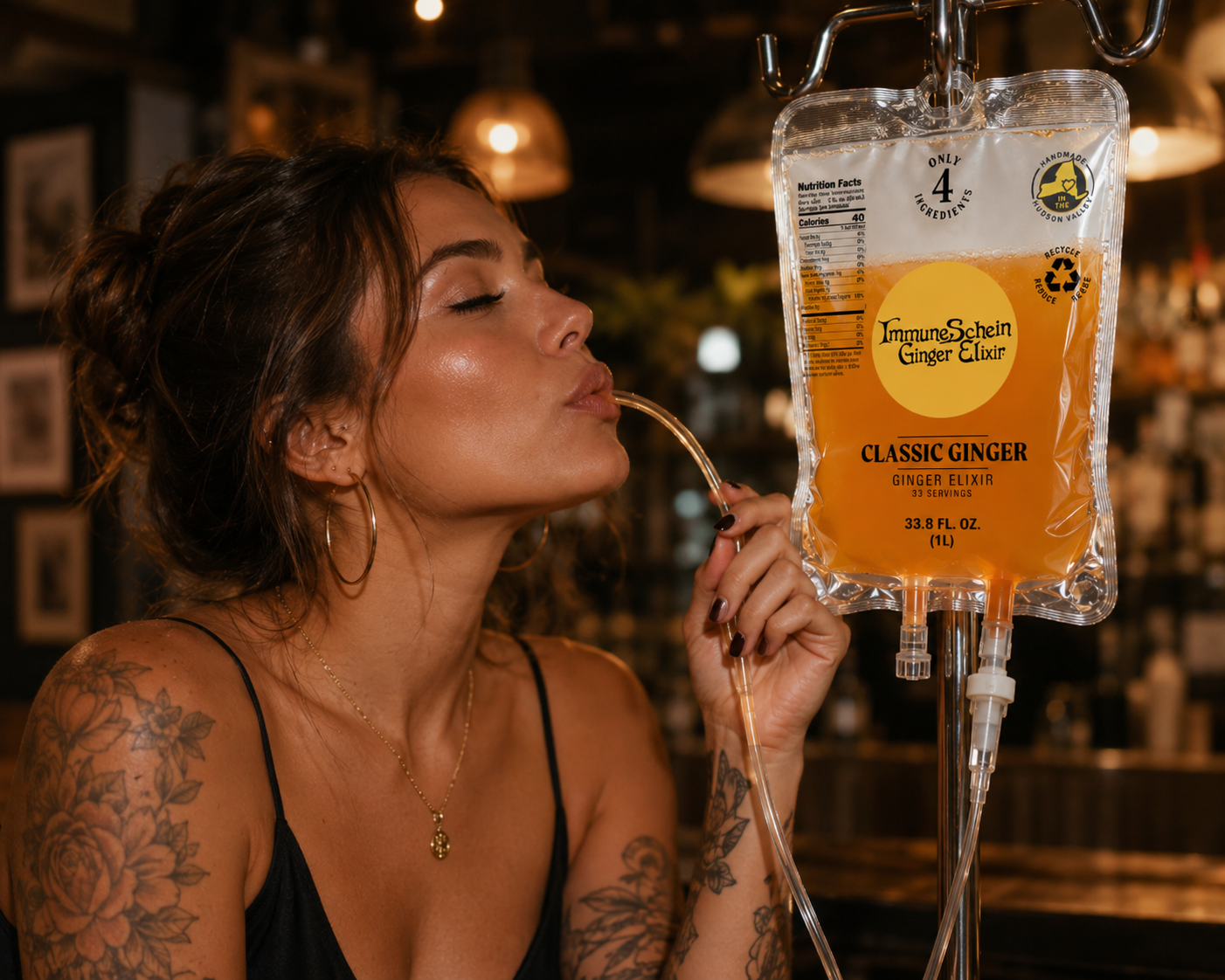 A woman with tattoos on her arms and shoulder, wearing large hoop earrings and a black top, is drinking ginger elixir from an IV bag labeled 'ImmuneSchein Ginger Elixir' labeled 'Classic Ginger' and containing orange liquid, in a bar or cafe setting.