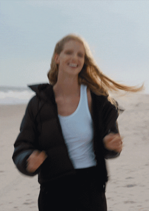 A woman running on the beach with a smile, wearing a black jacket and a white tank top.