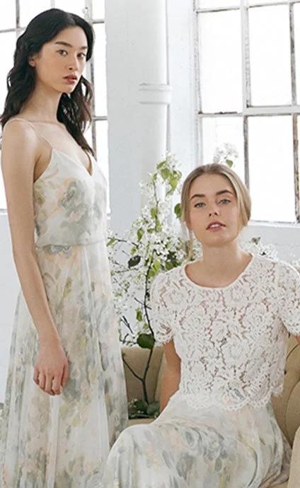 Two women wearing light-colored, floral-patterned dresses in a bright room with large windows and white walls, surrounded by plants and flowers.