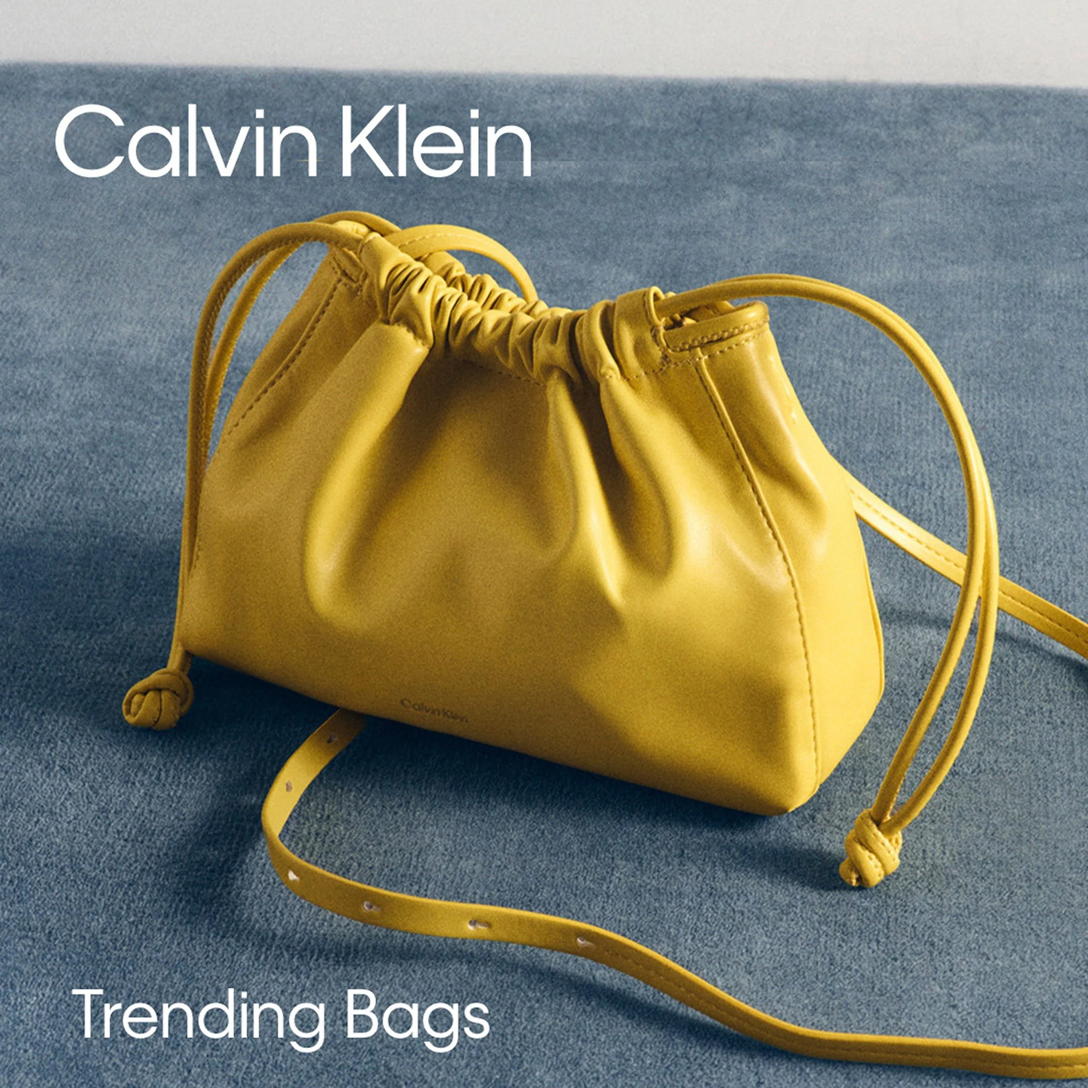 Yellow Calvin Klein trending bag with attached matching strap on a blue textured surface.