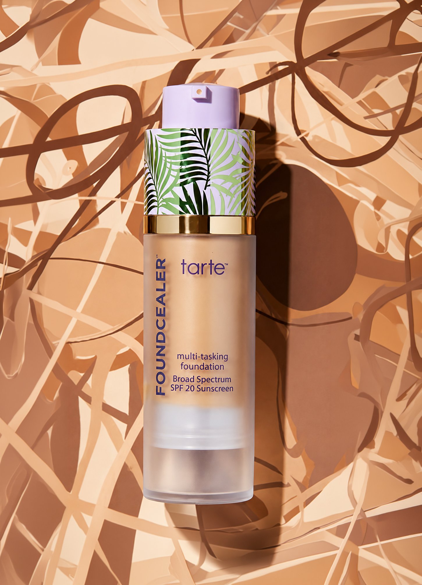 A bottle of Tarte foundation with a tropical leaf pattern on the cap, placed on a beige and brown abstract background.
