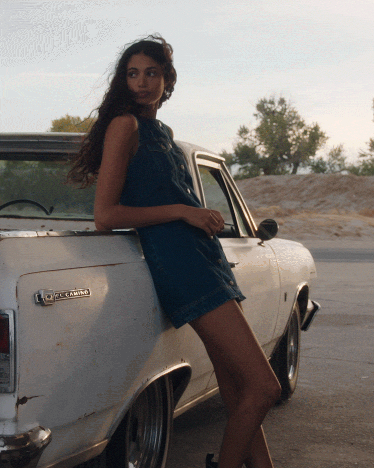 A young woman leaning against the side of an old white El Camino truck, wearing a denim dress, with trees and a cloudy sky in the background.