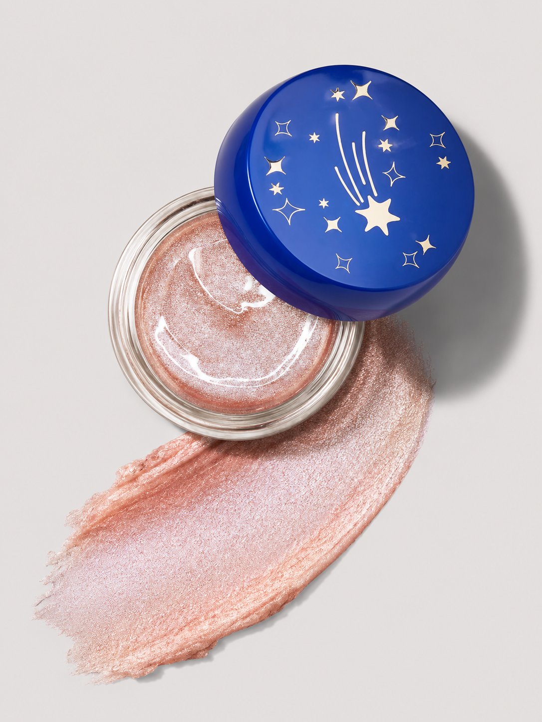 Open jar of shimmery, pink cosmetic or skincare product with a lunar and star design on the blue lid, and a swipe of the product on a white surface.