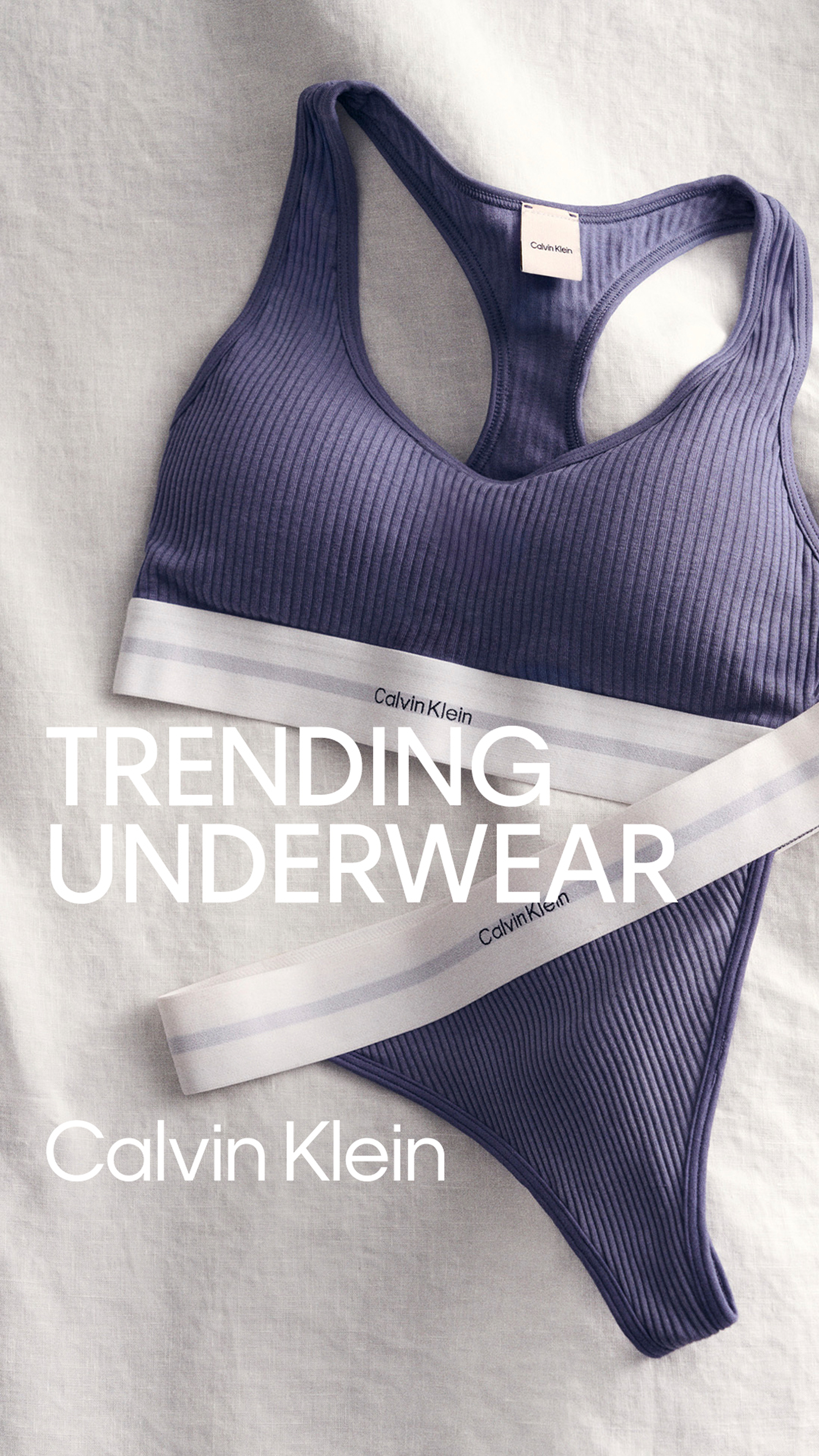 A purple Calvin Klein sports bra with a ribbed texture, laid on a white surface. The bra has a white elastic band with gray stripes and the Calvin Klein logo, with text overlay reading "Trending Underwear" and "Calvin Klein."