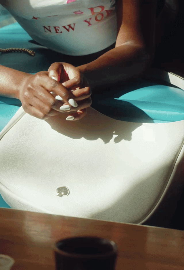 Close-up of a woman with manicured nails, clasping her hands on a white table with a small silver heart pin, wearing a black top and a shirt that says 'New York.'