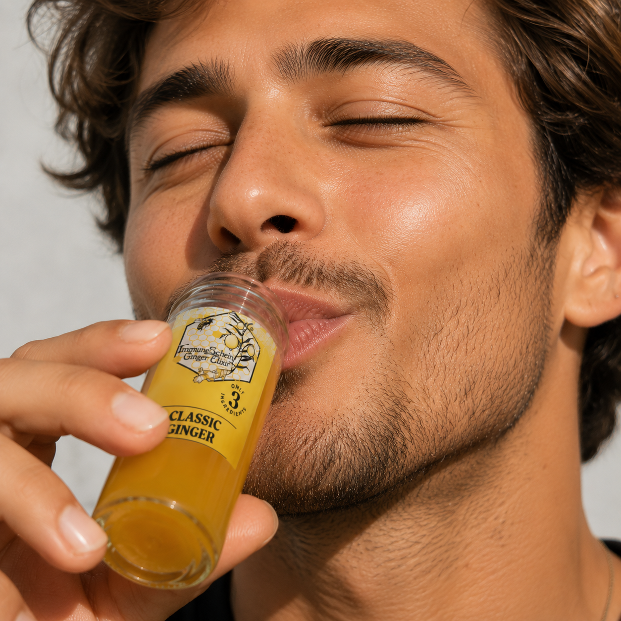 A man with closed eyes and facial hair drinks ginger elixir from a small glass with a yellow label reading "Ginger Elixir" and "Classic Ginger".