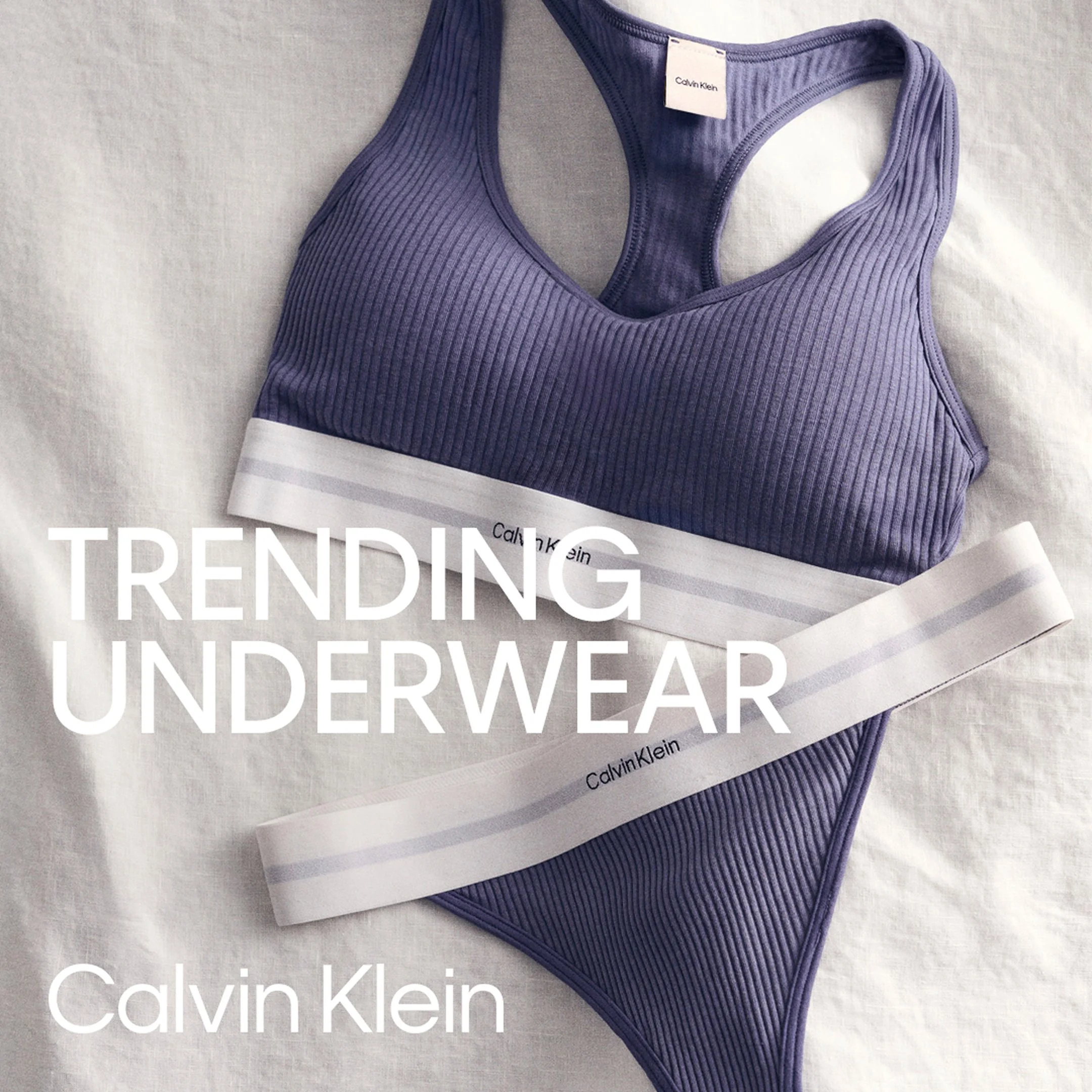 A purple Calvin Klein sports bra with a racerback design and a white band at the bottom, placed on a light-colored fabric background with a Calvin Klein magazine overlay.