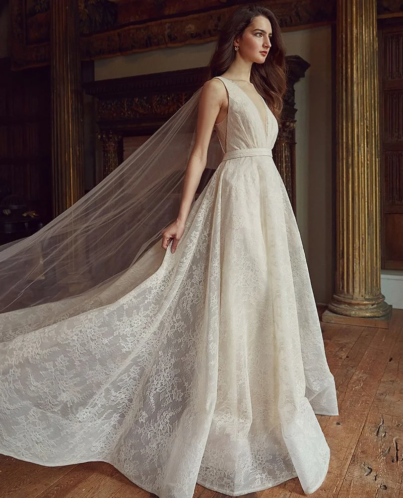 A woman in a white wedding dress with a long train and lace details, standing inside a wooden room with ornate columns.