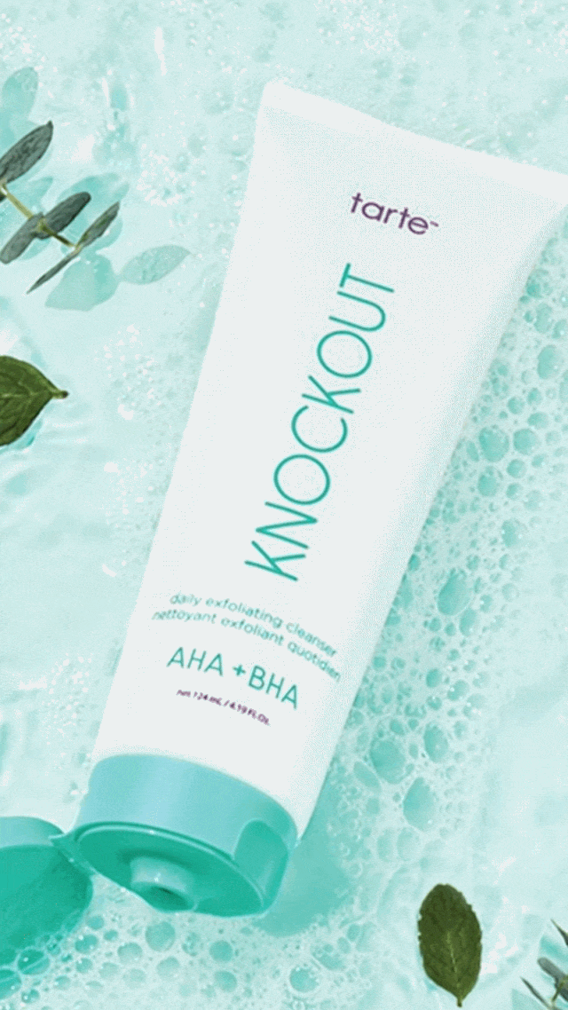 A white tube of Tarte Knockout daily exfoliating cleanser with AHA and BHA, surrounded by bubbles and green leaves.