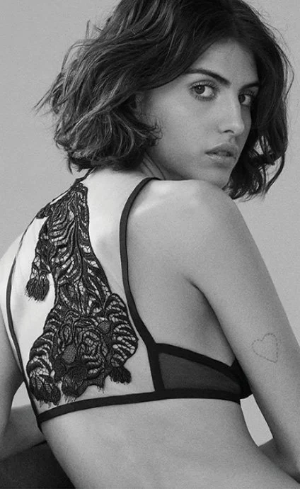 Black and white photo of a young woman with shoulder-length wavy hair, wearing a lace bralette, looking over her shoulder.