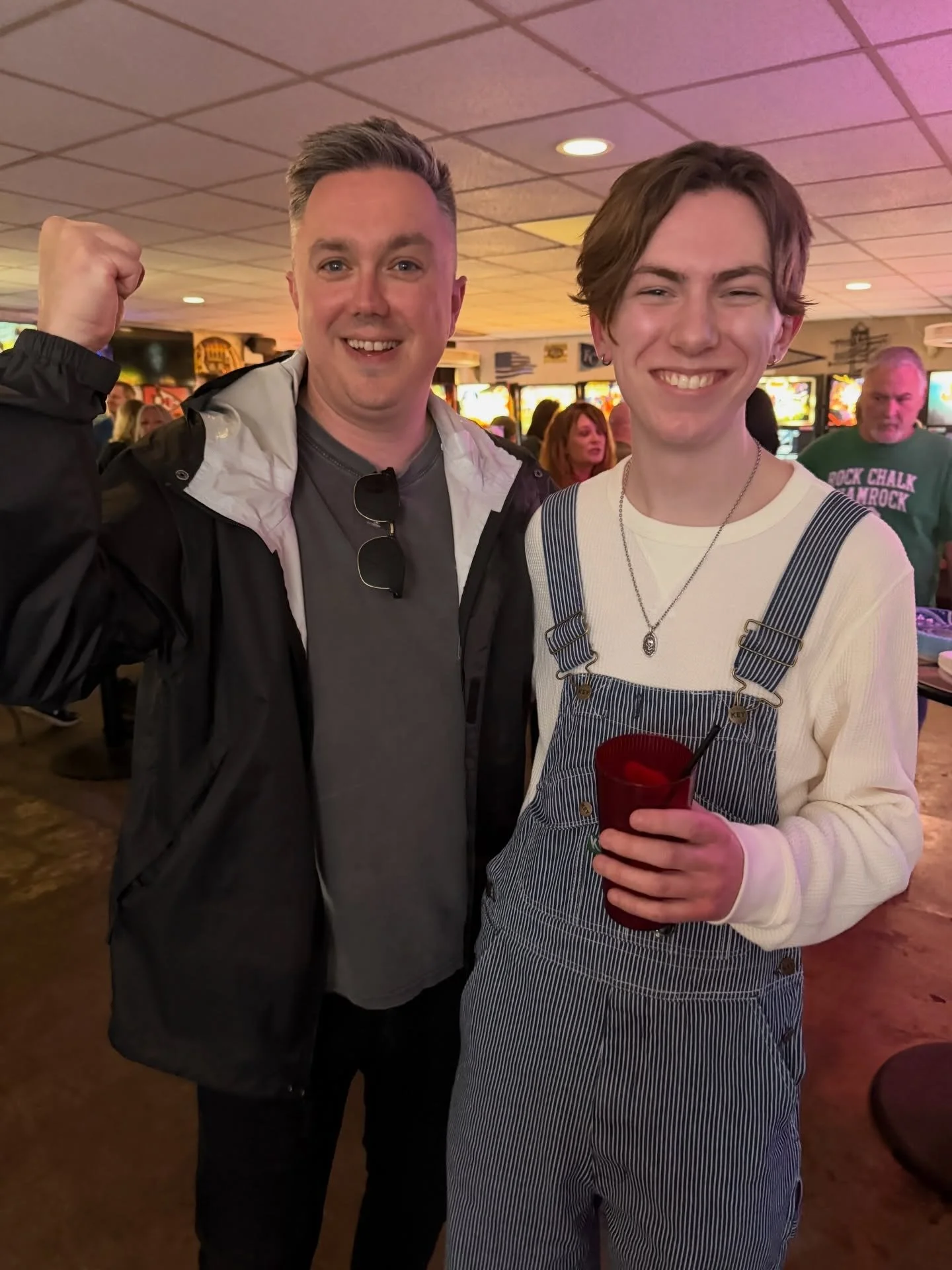 Two young men smiling indoors at a bar or pub, one with a raised fist and the other holding a red cup.
