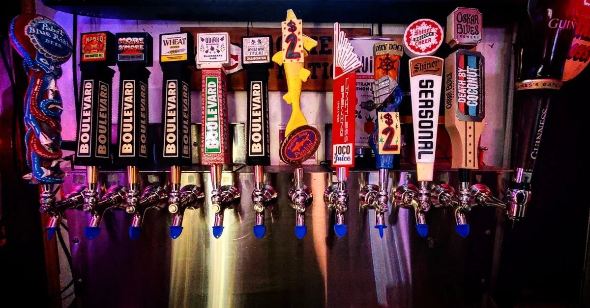 Multiple beer taps with colorful labels in a bar, some with price signs and decorative items on top.