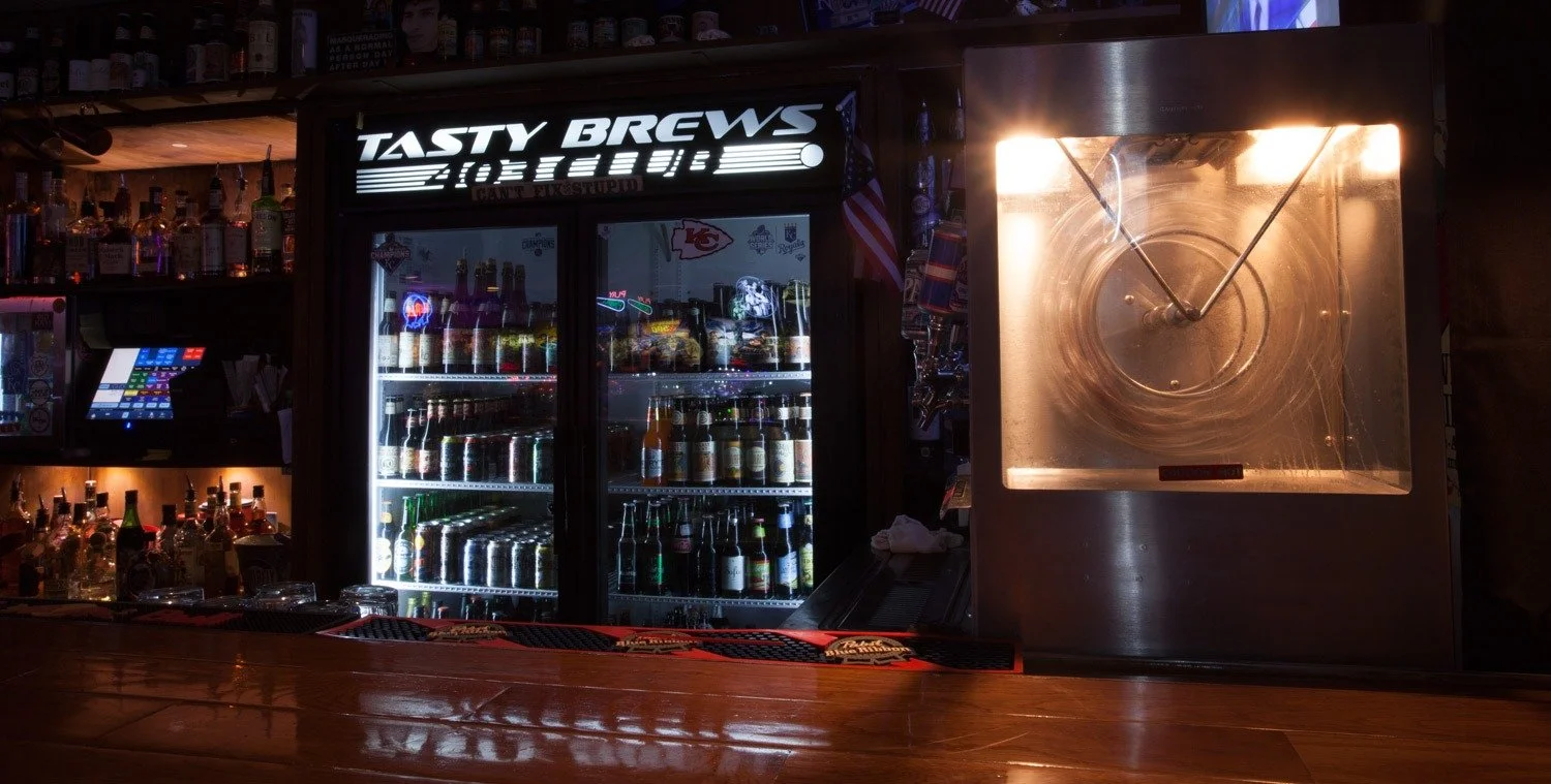 Bar scene with a fridge full of bottles and cans, a drink dispensing machine on the right, and a neon sign that reads 'Tasty Brews.'