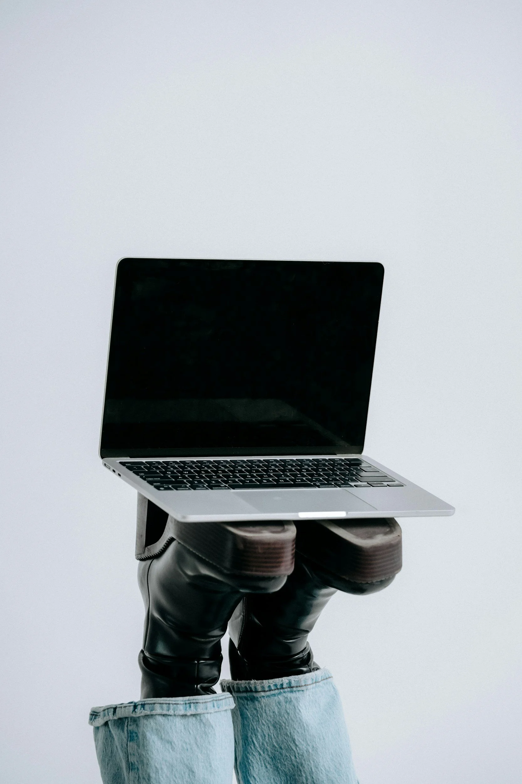 Person holding a laptop with the screen facing outward, wearing black gloves, light blue jeans with cuffs rolled up, and black shoes, against a plain gray background.