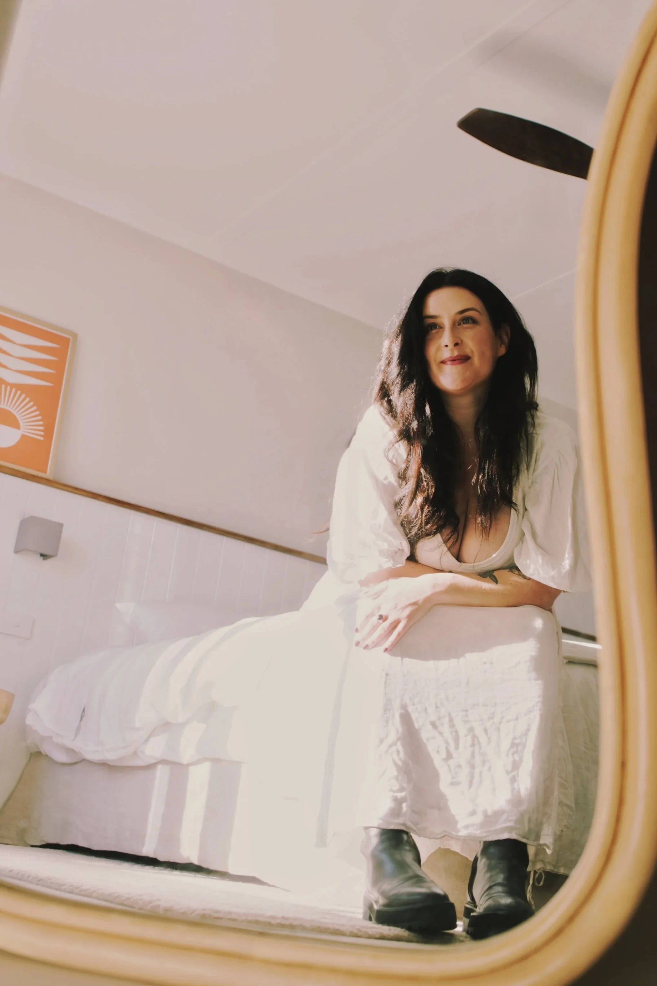 A woman with long dark hair, smiling and wearing a white dress, is sitting on a bed and looking at her reflection in a mirror. She is wearing black boots, and the room has light-colored walls with a framed artwork on the wall.