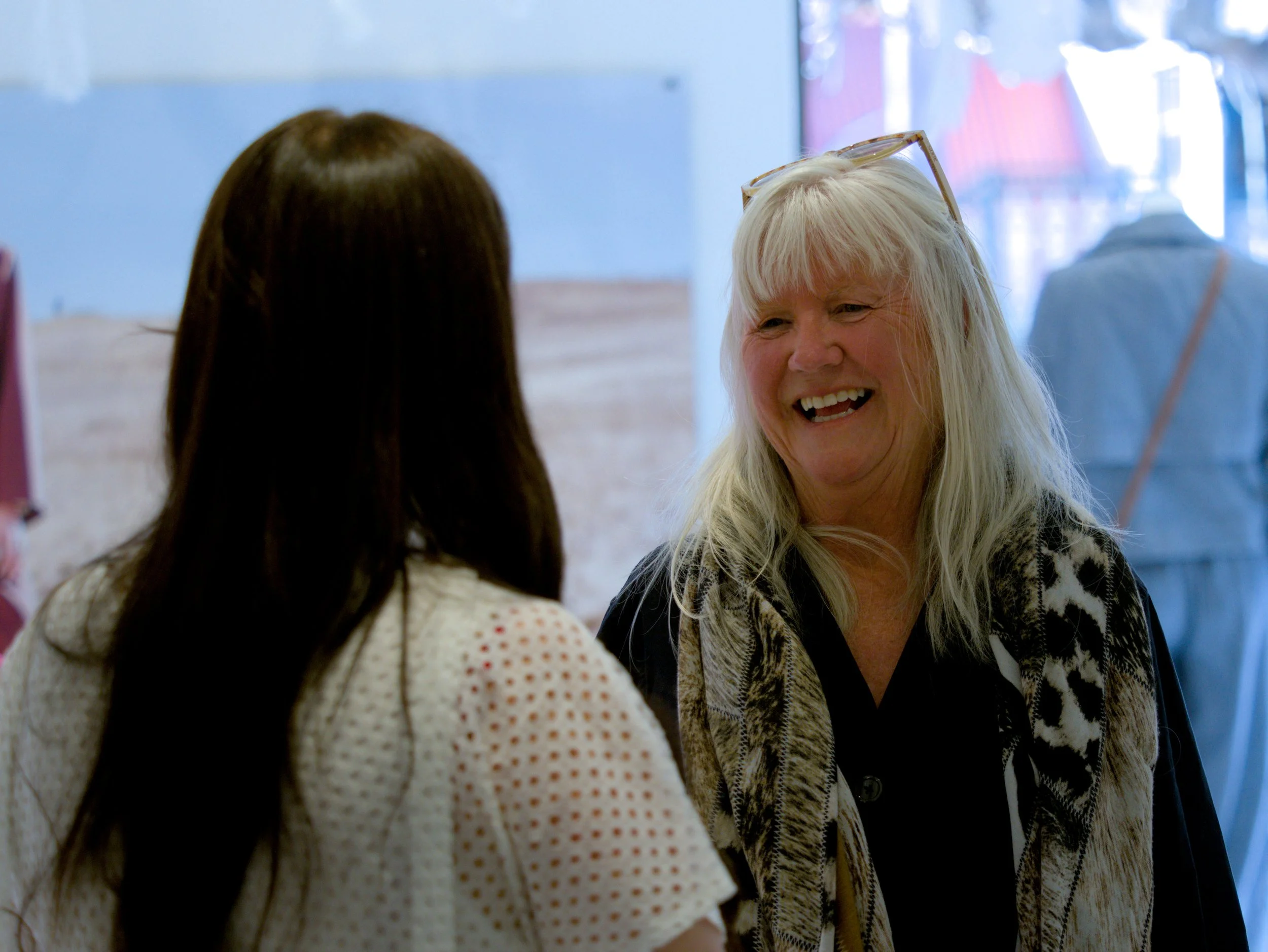 Two women talking in a bright indoor space, one with blonde hair and glasses on her head smiling, and the other with dark hair seen from behind.