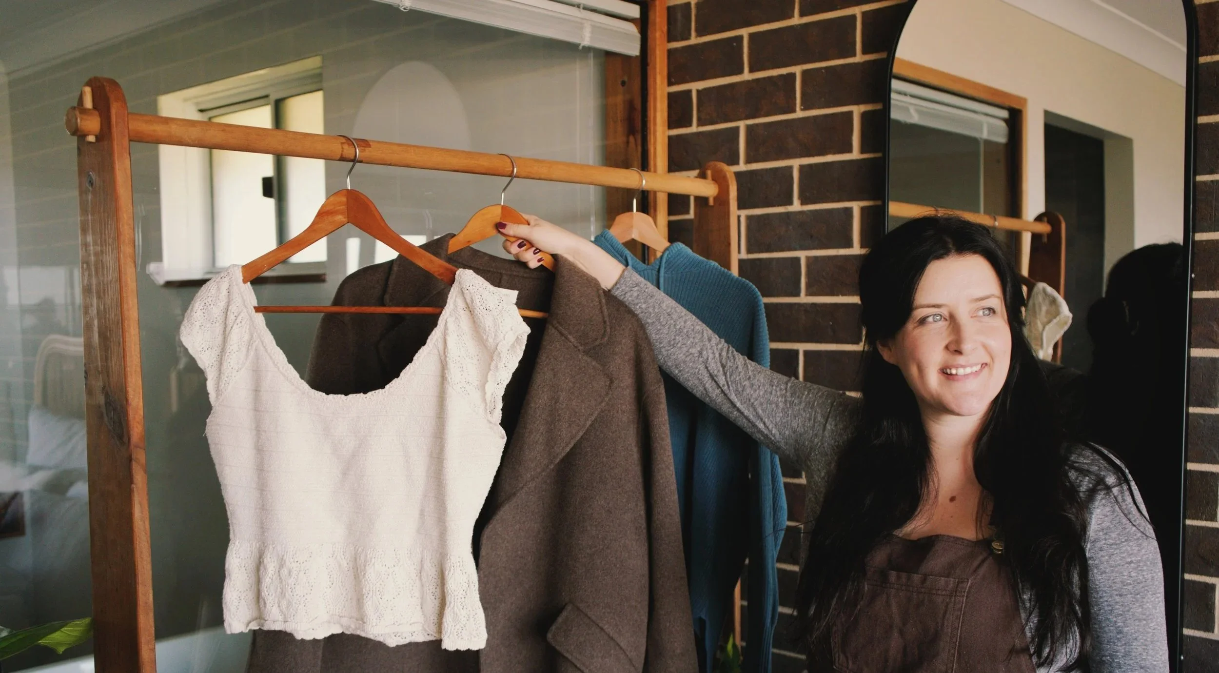 Woman with black hair smiling as she looks into a mirror, with hanging clothes including a white knitted top and a brown jacket on a wooden clothing rack in a room with brick and siding walls.