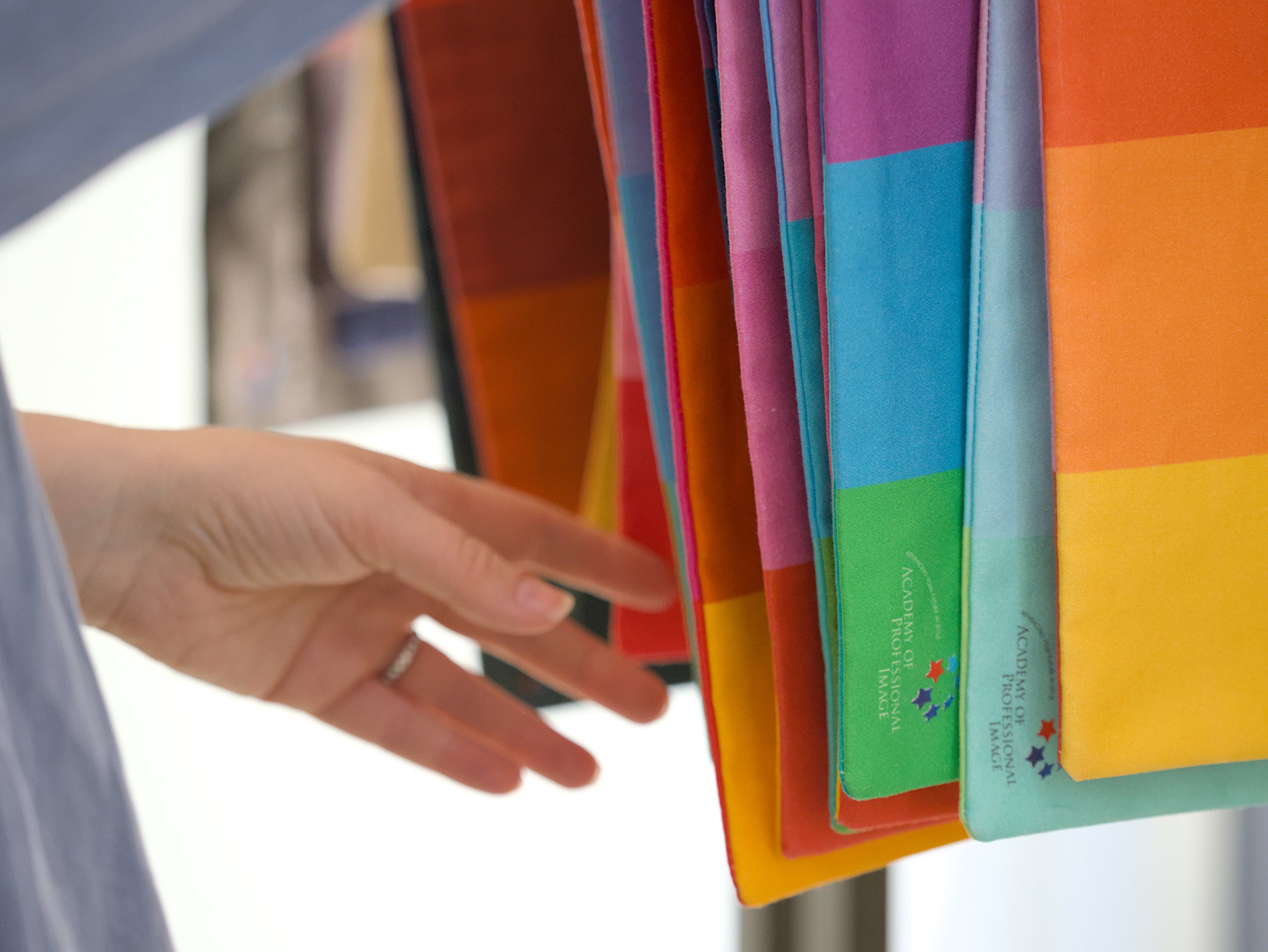 Close-up of a person's hand sorting colourful fabric drapes with text on their tabs, displayed vertically with a variety of colors such as yellow, orange, pink, blue, green, and purple.