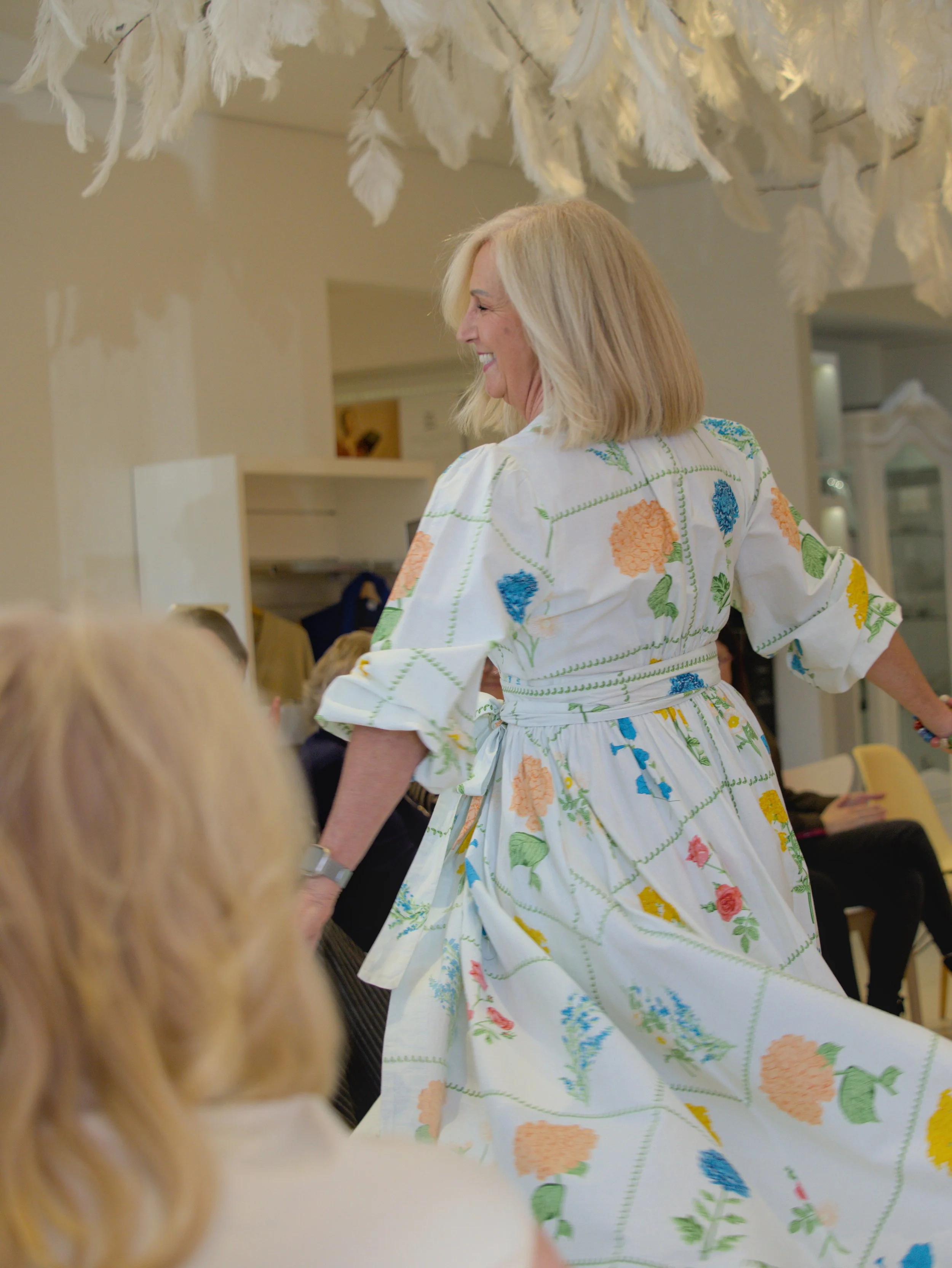 A woman with blonde hair smiling and wearing a white dress with colorful floral patterns, interacting with people in an indoor setting.