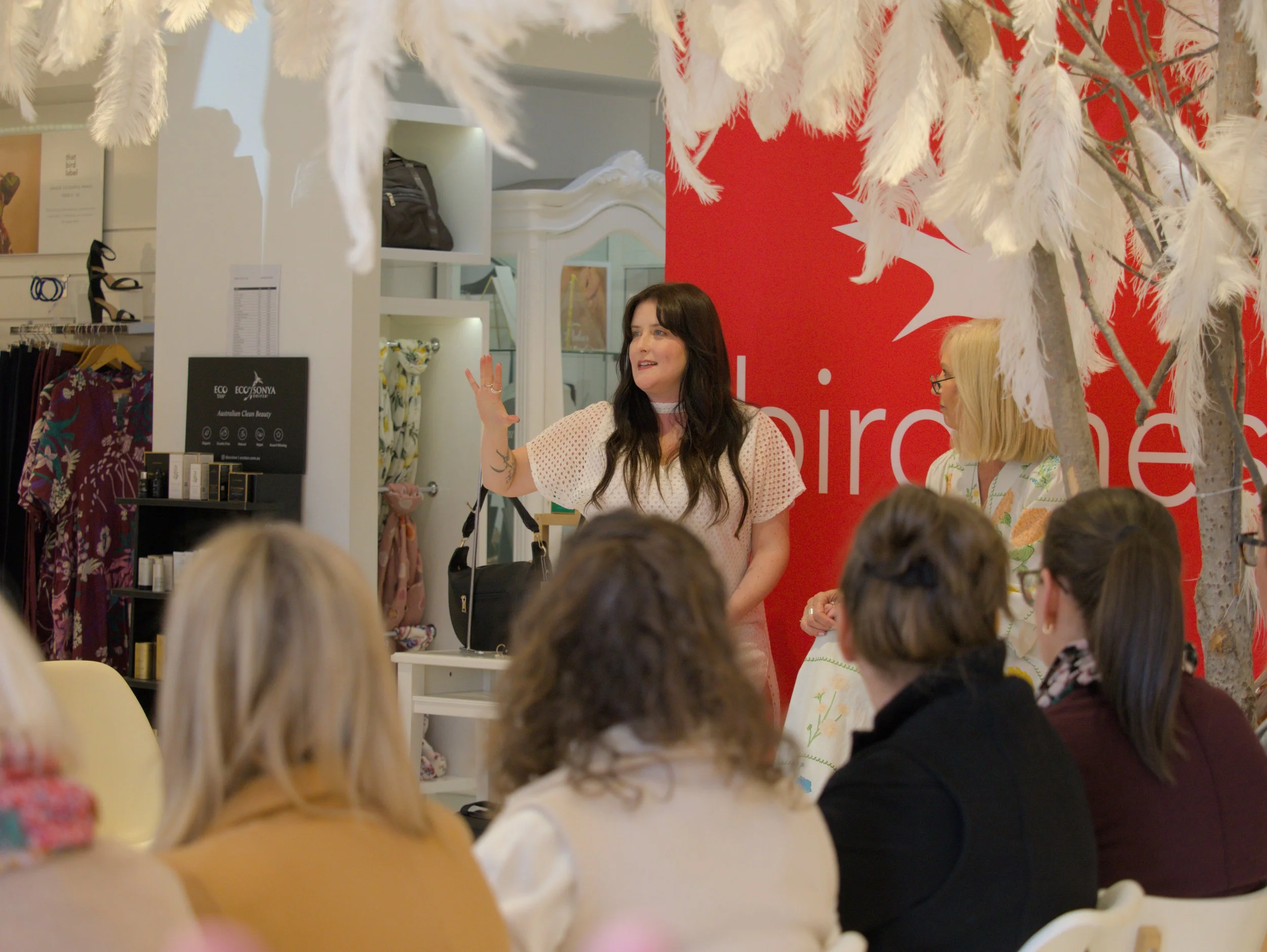Two women speaking to an audience in a retail space, surrounded by customers and display shelves.