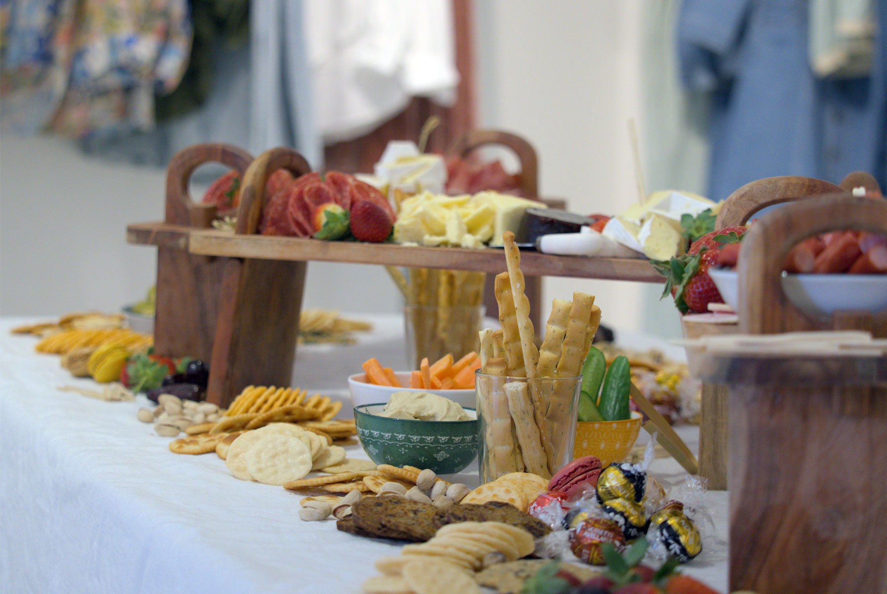 An assortment of cheeses, fruits, crackers, and snacks displayed on a table with a white tablecloth, featuring a wooden serving stand and colorful bowls.