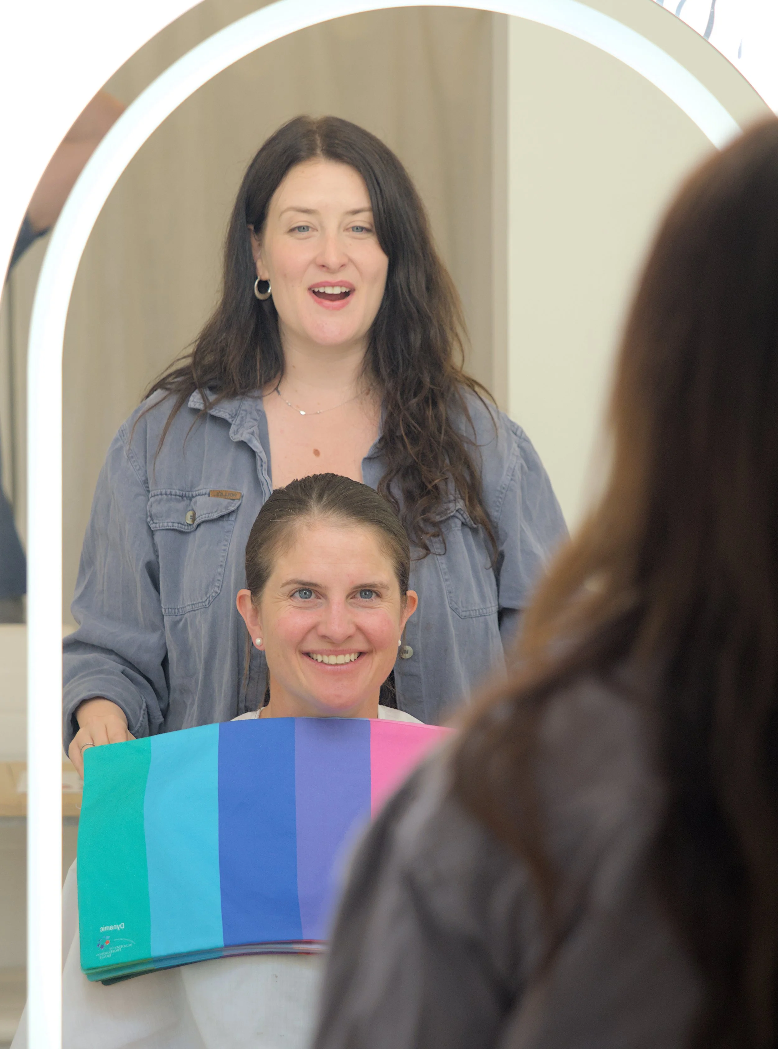 A woman with dark hair and earrings looking into a mirror, smiling. She is standing behind a person holding a colour analysis diagnostic drape, partially visible in the foreground. The setting appears to be indoors.