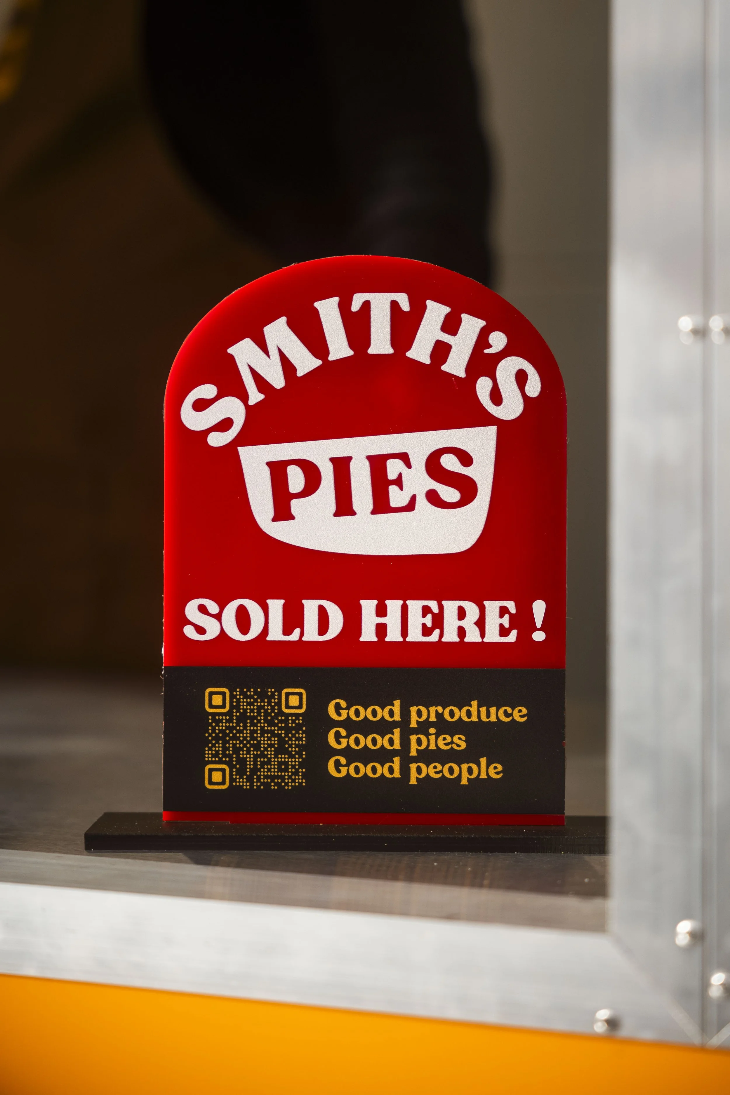 Red sign with white text reading "SMITH'S PIES SOLD HERE!" and yellow text that says "Good produce, Good pies, Good people" with two QR codes at the bottom.