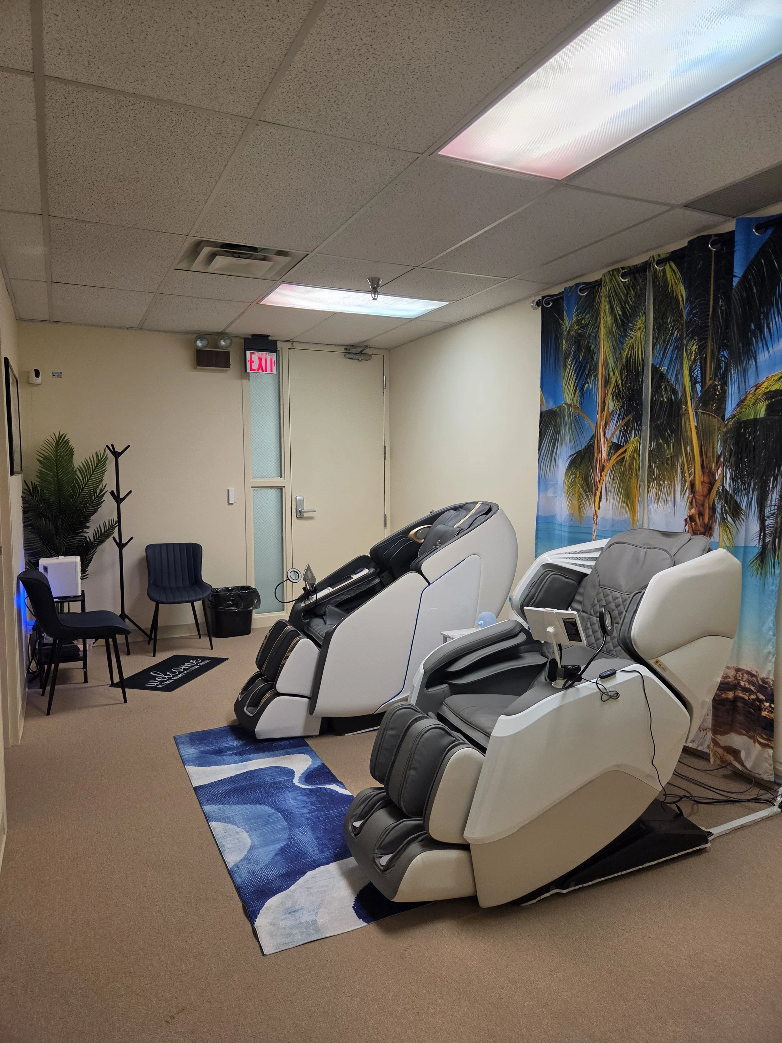 Massage chairs in a spa or massage therapy room with a tropical beach backdrop curtain, chairs, a small table with a device, and chairs along the wall.