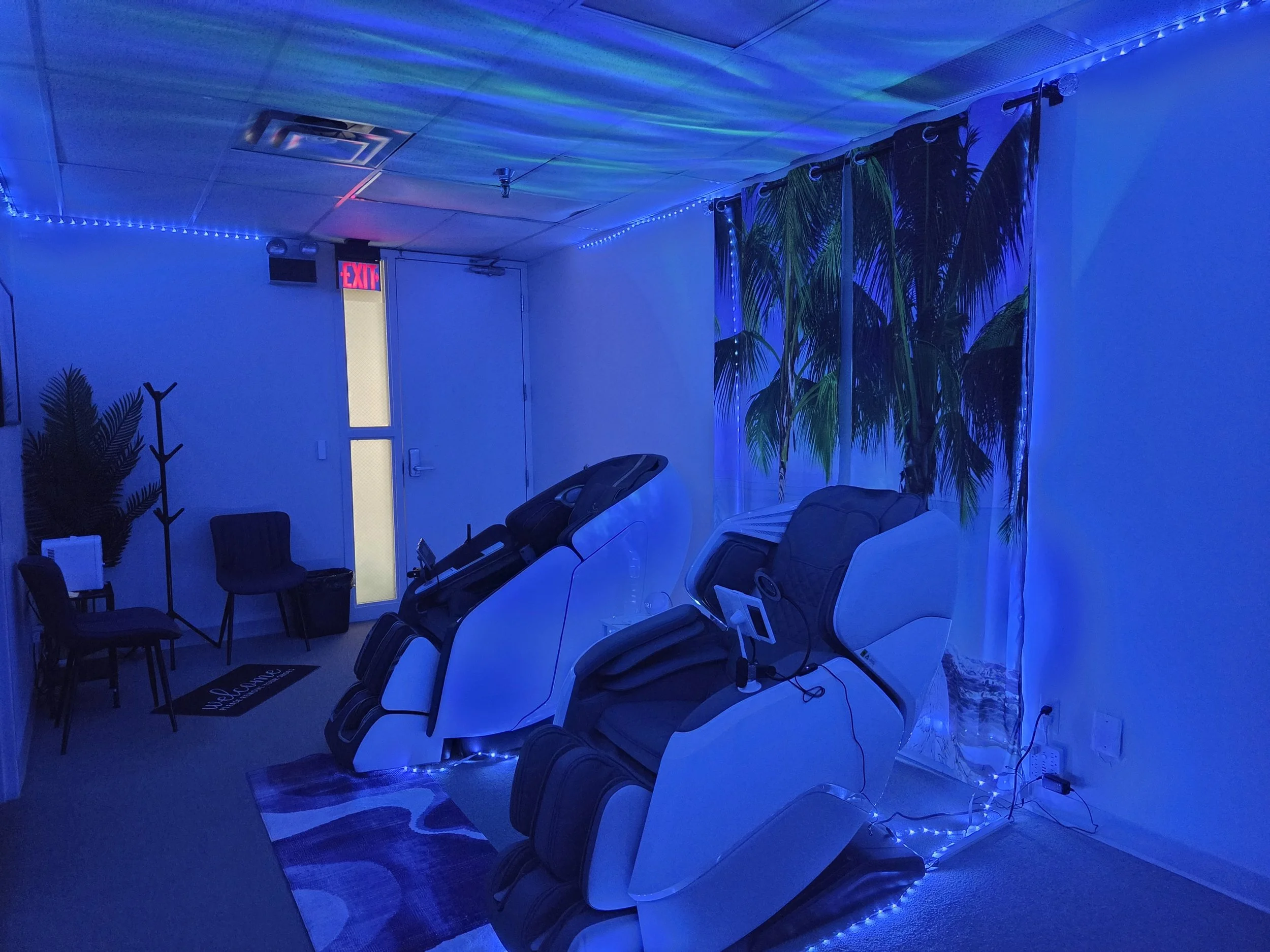 A room with blue lighting, featuring a row of massage chairs, a large plant, and a door with an illuminated 'EXIT' sign.