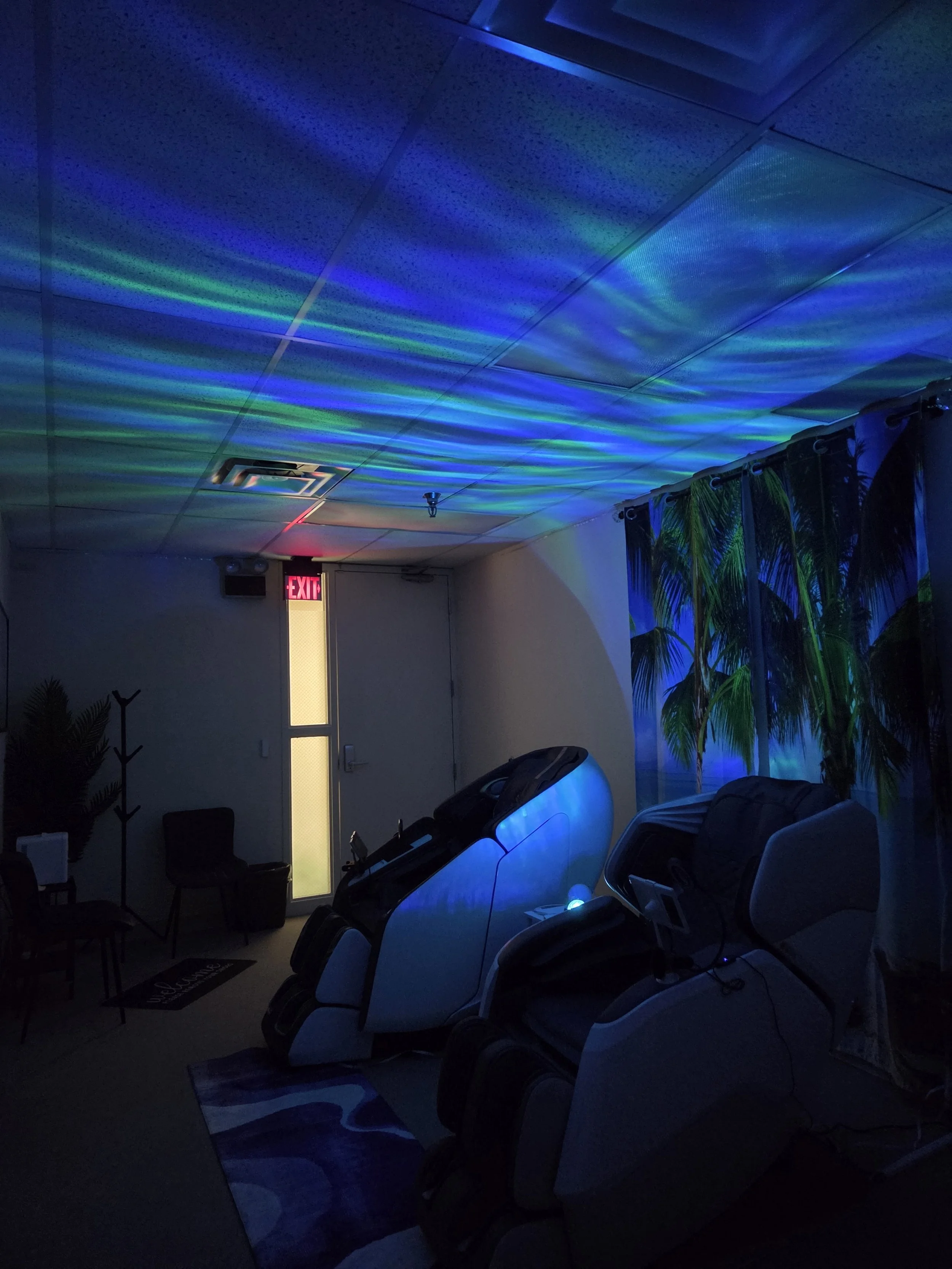 Indoor room with massage chairs, rainbow-colored light projections on the ceiling, potted plant and a decorative curtain of palm leaves, illuminated exit sign, dark ambiance